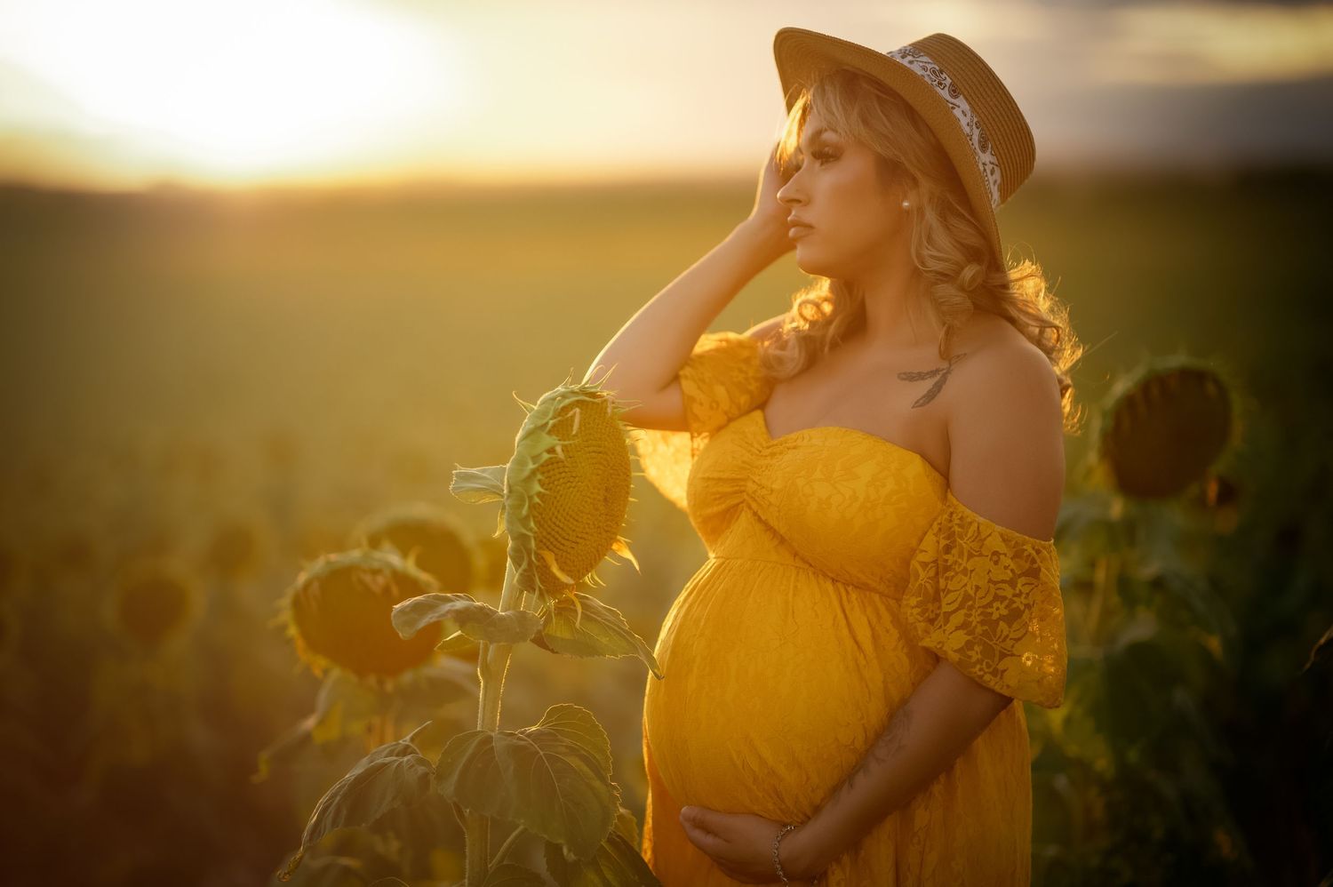 Pregnant woman in yellow dress holding belly in sunflower field at golden sunset.