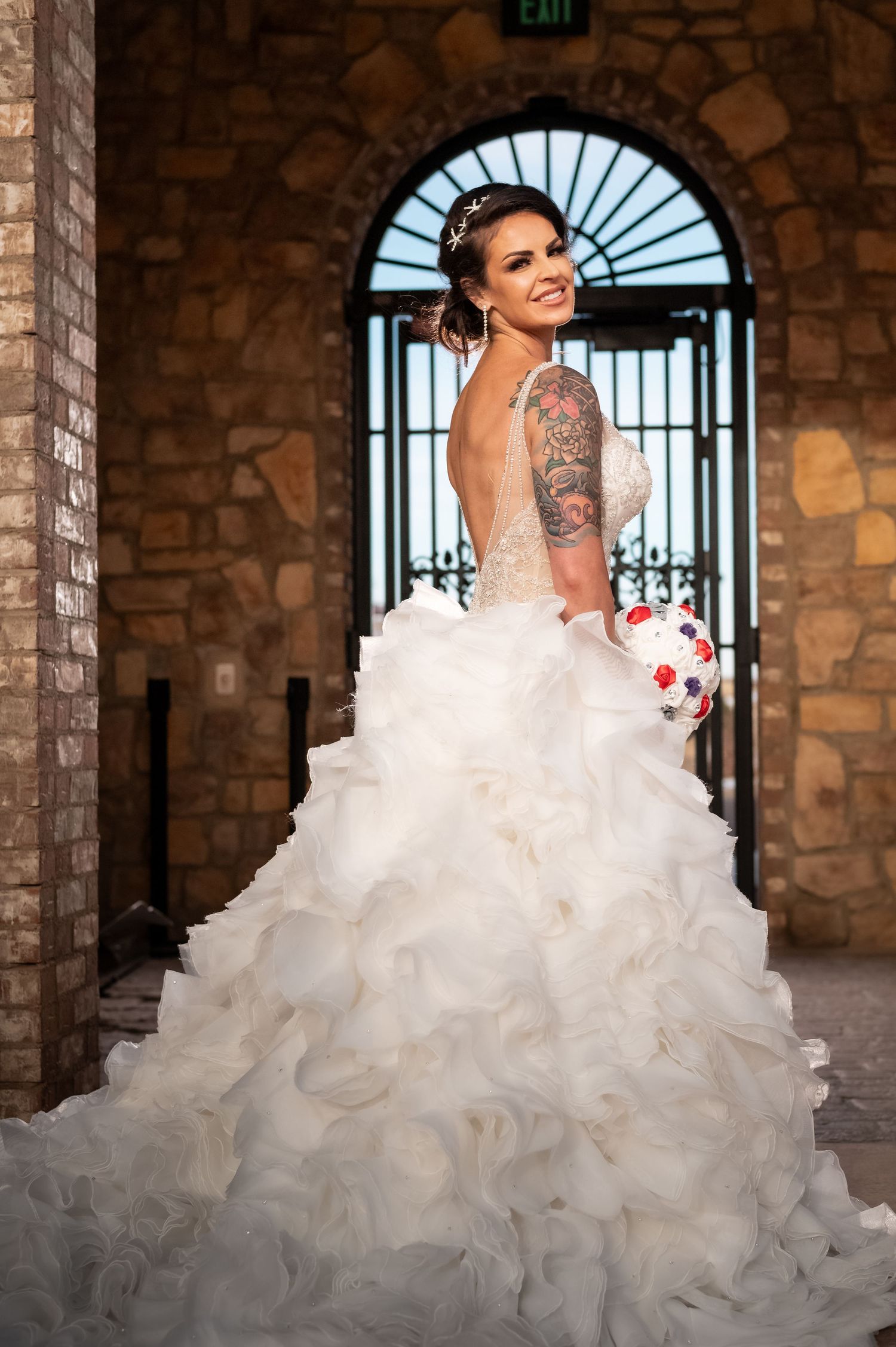 Bride in stunning backless ruffled ball gown with tattoos holding bouquet in stone archway.