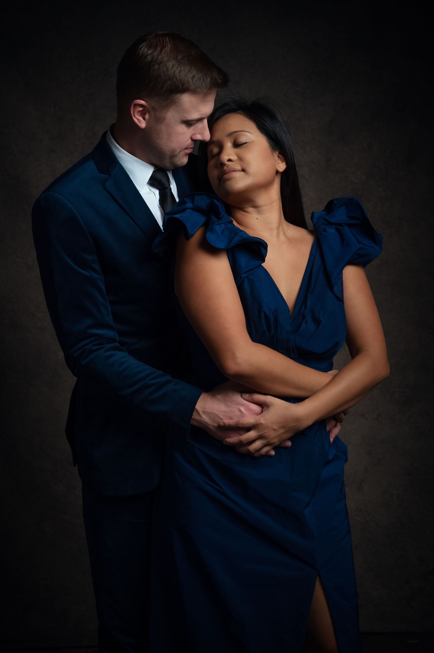 Elegant couple in matching blue outfits embracing in dramatic studio lighting, man in suit holding woman from behind.