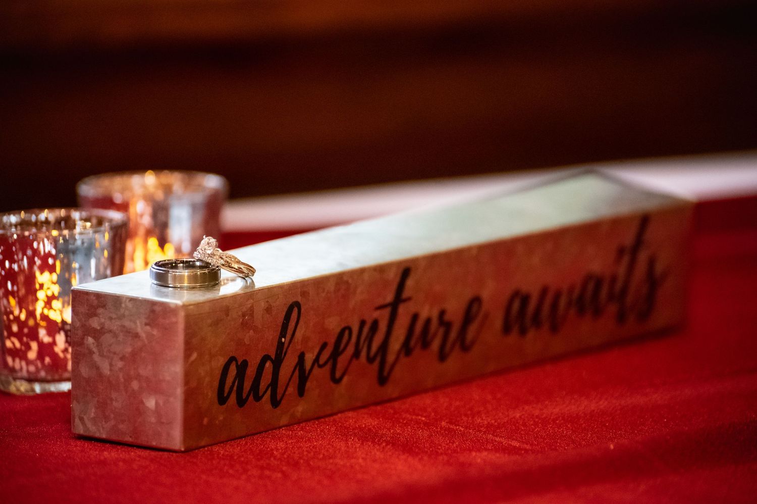 Wooden wedding ring box engraved with adventure awaits script, holding gold rings beside red glitter votive candles.