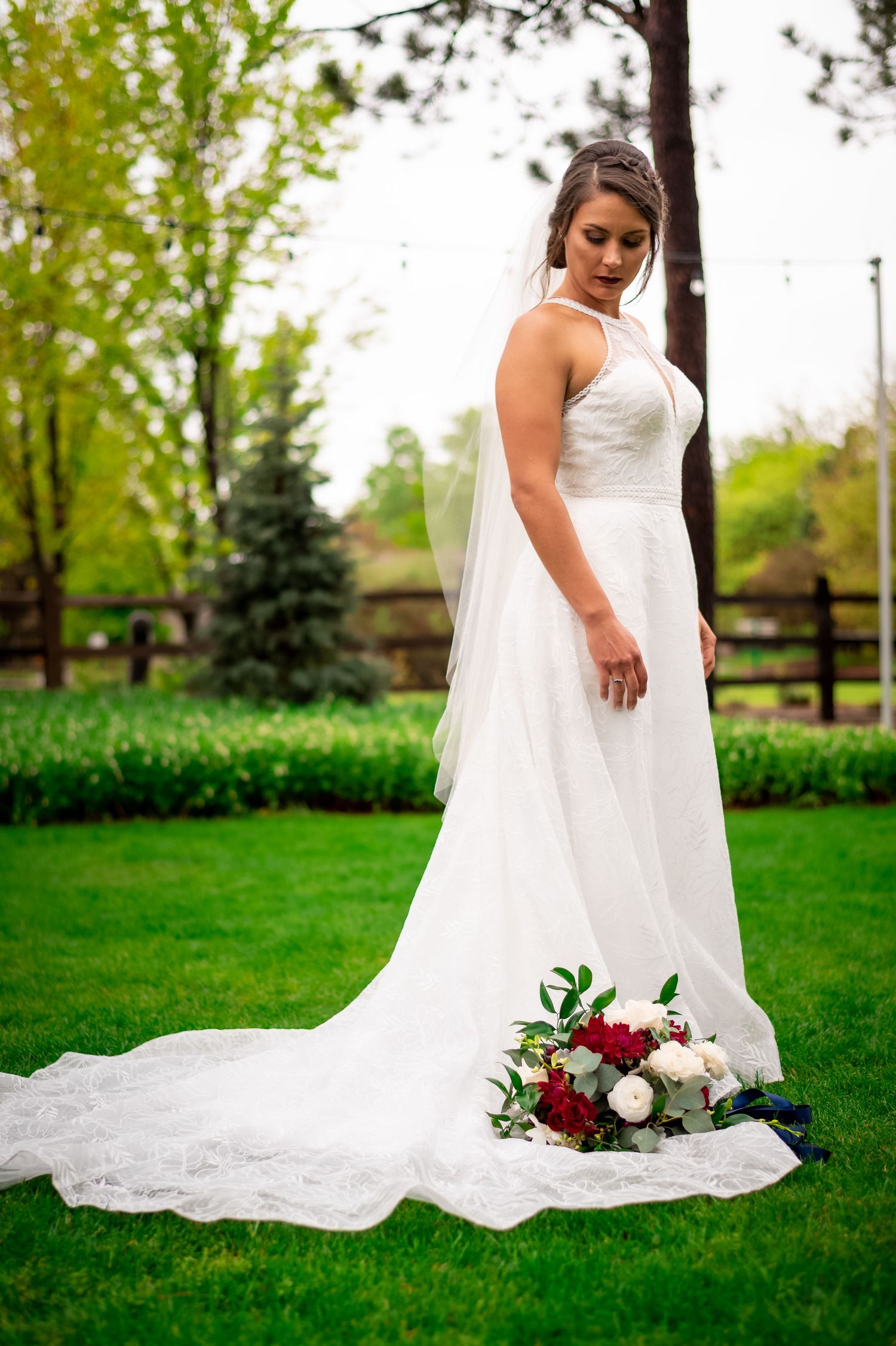 Bride in halter wedding gown with long train stands on green lawn.