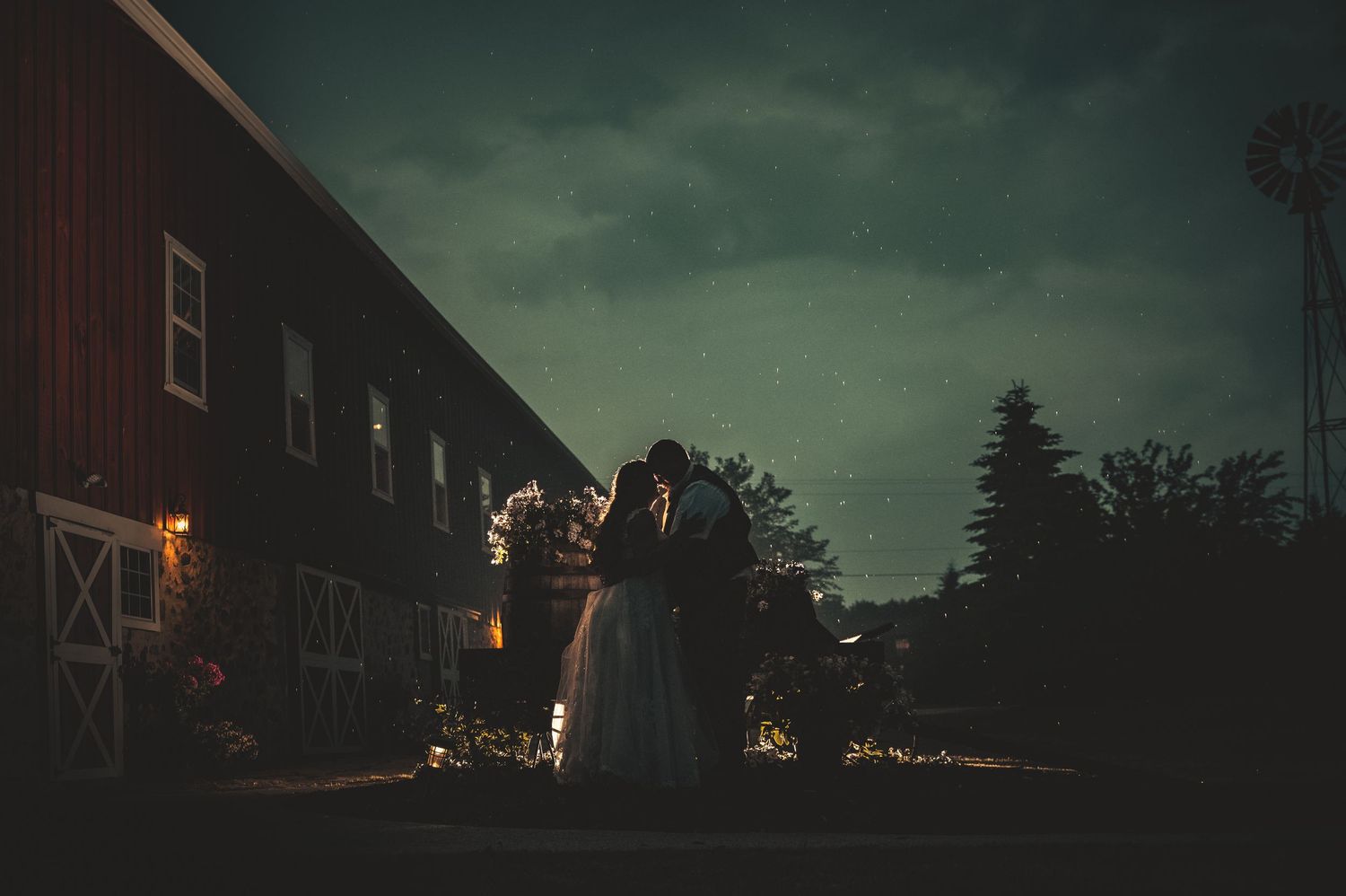 Silhouette of bride and groom kissing outside red barn at twilight.