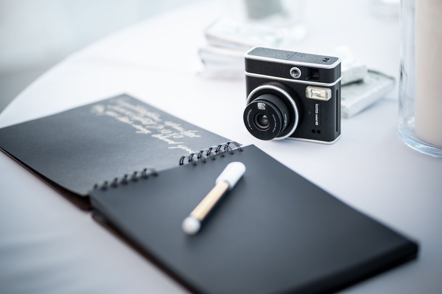 Instax mini camera beside black guest book and pen on white table.