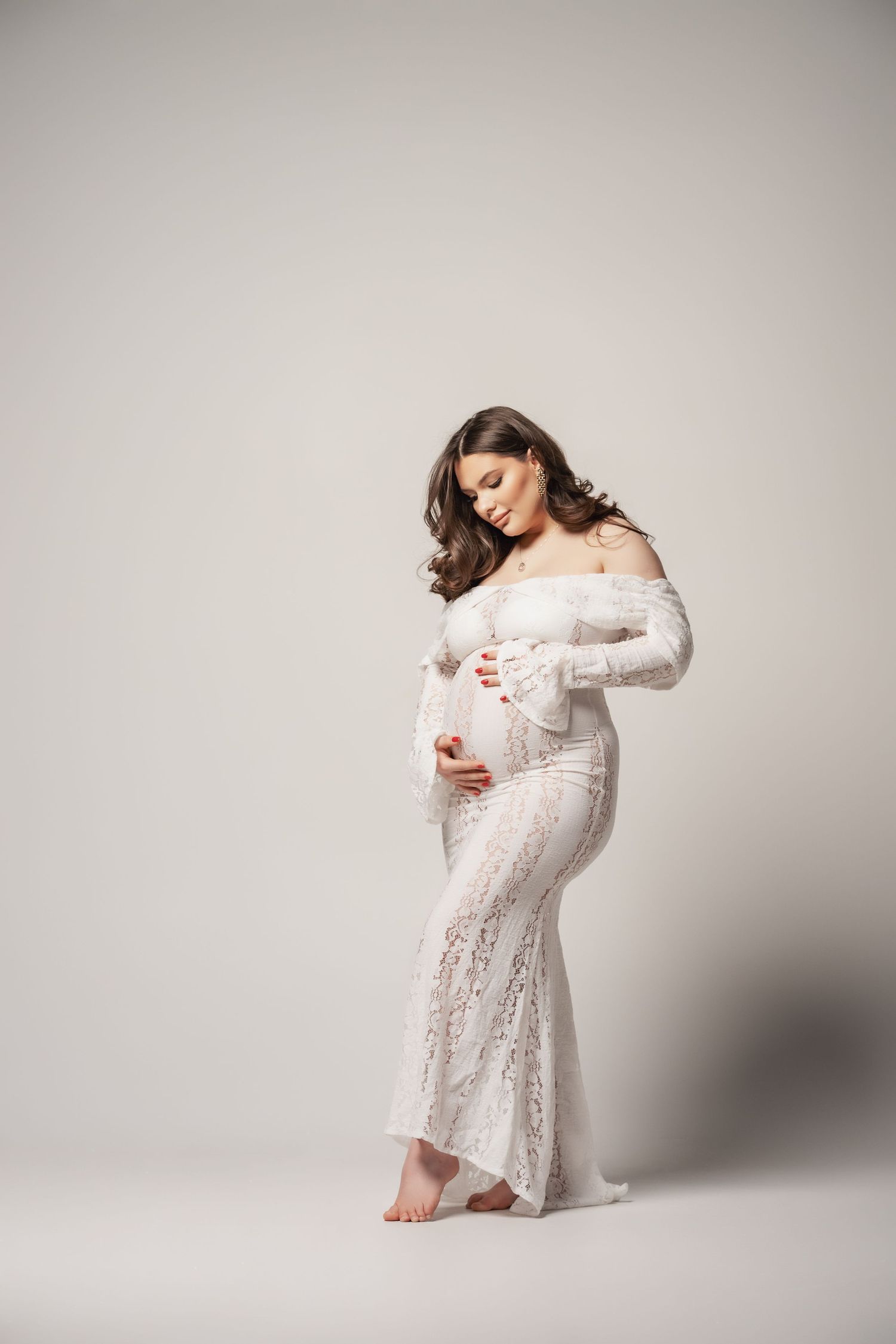 Pregnant woman in elegant white lace off-shoulder gown cradling her baby bump in a soft studio setting.