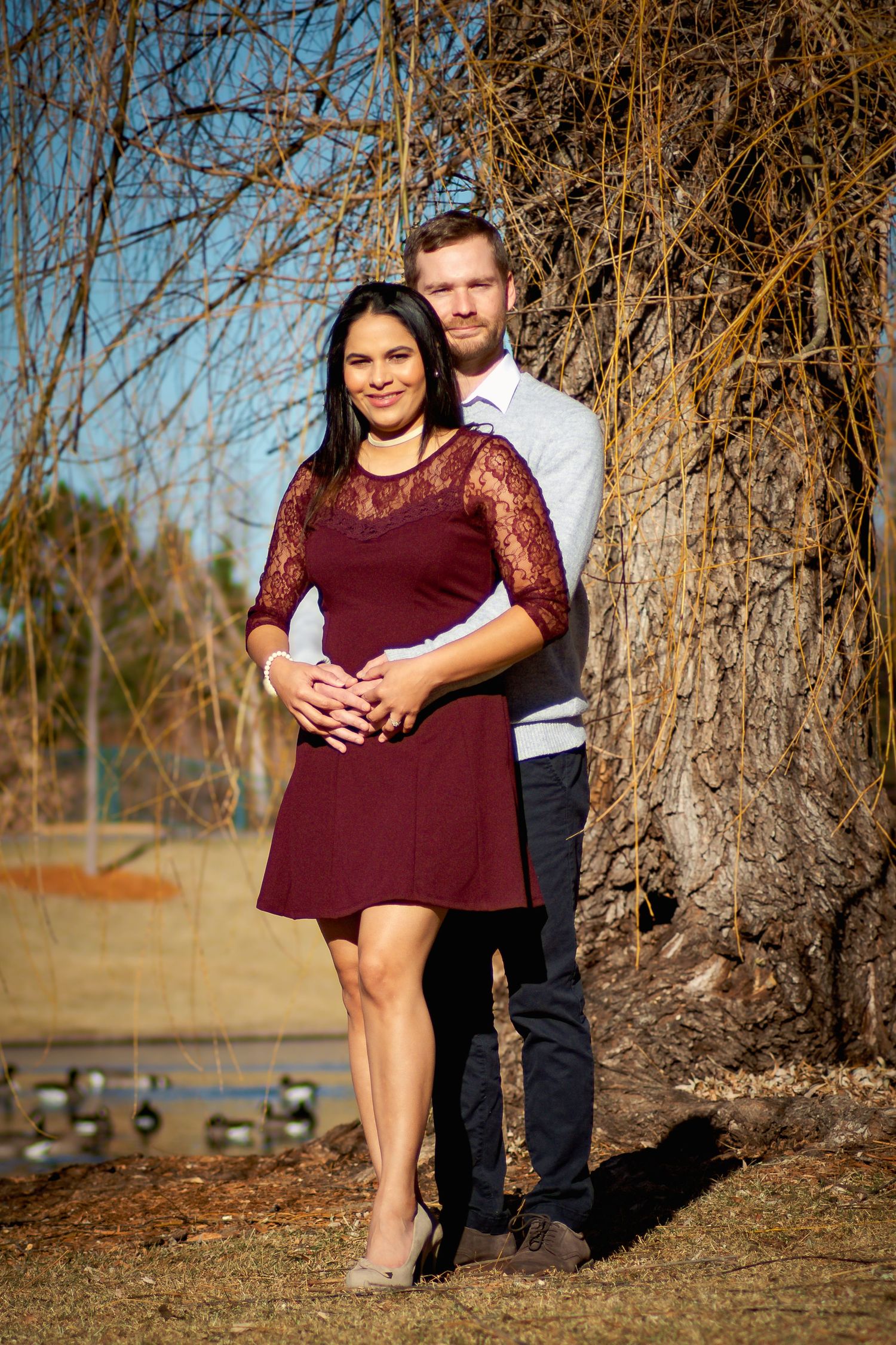 Couple posing together near a bare tree outdoors, woman in burgundy dress with man behind her embracing.