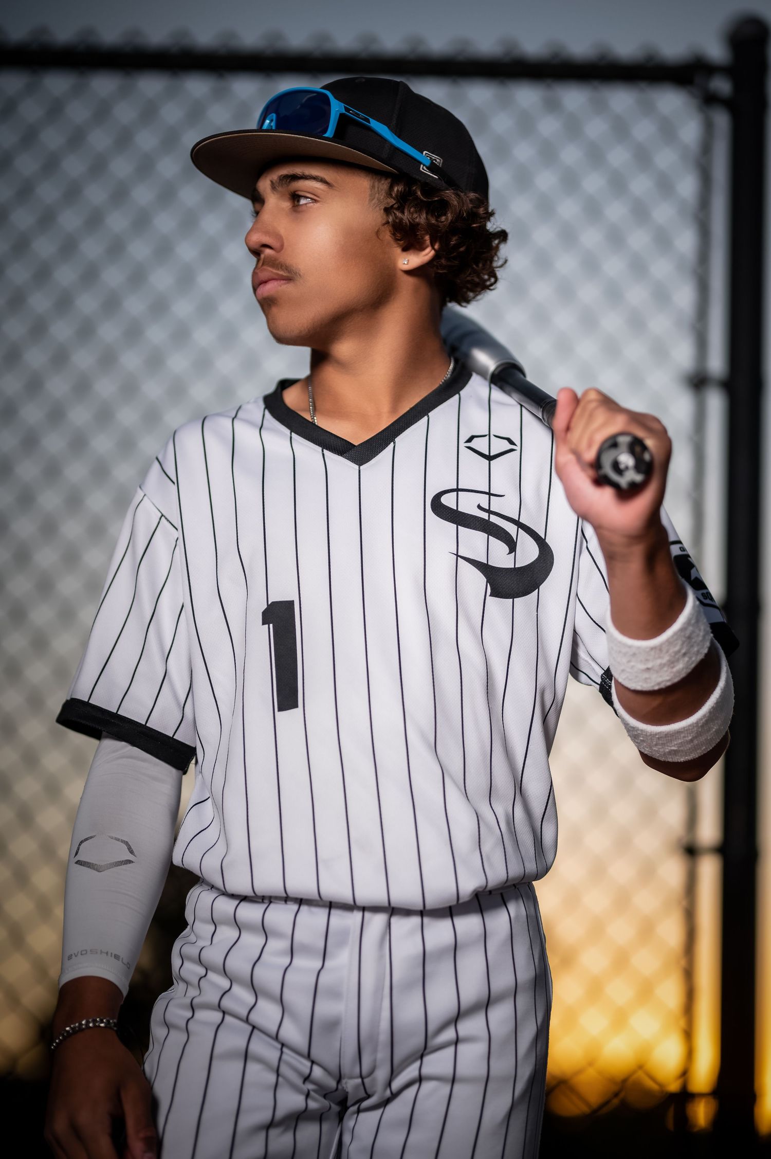 Young male baseball player in pinstripe uniform holding bat over shoulder near chain-link fence at dusk.