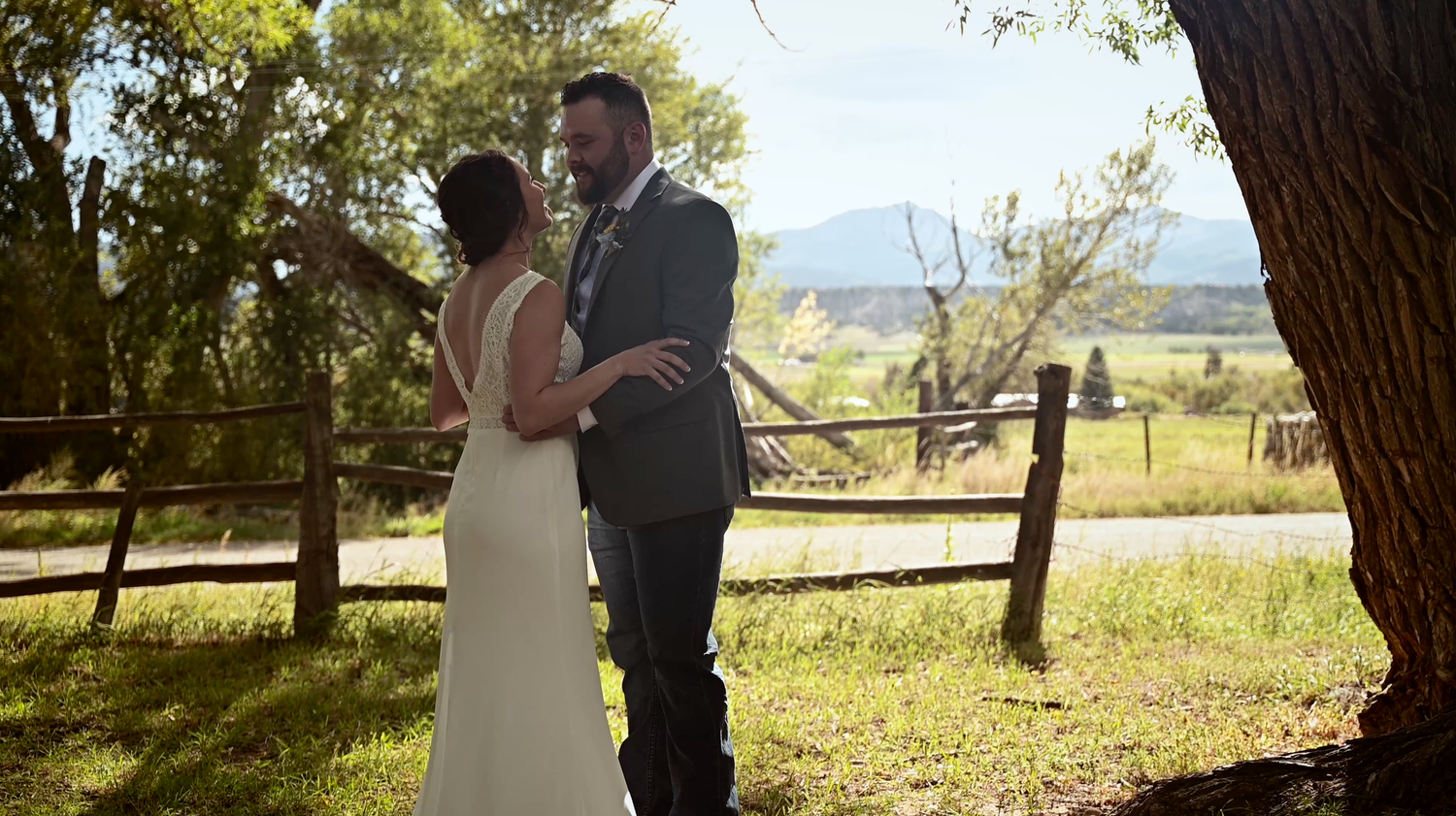 A bride and groom share a romantic outdoor moment near a rustic wooden fence surrounded by lush greenery.