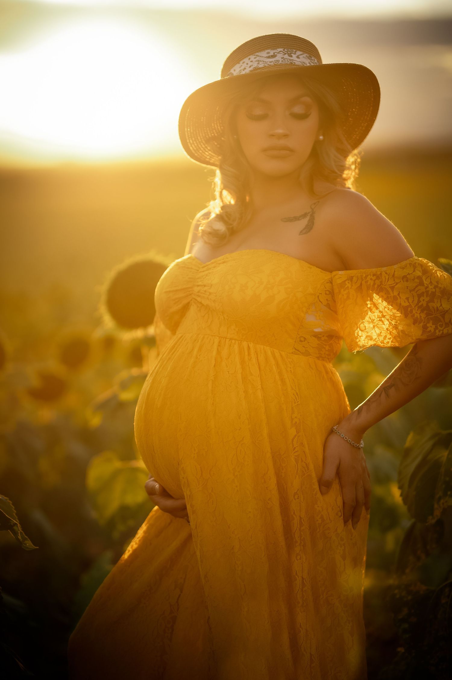 Pregnant woman in yellow lace gown and hat stands in sunflower field at golden sunset.