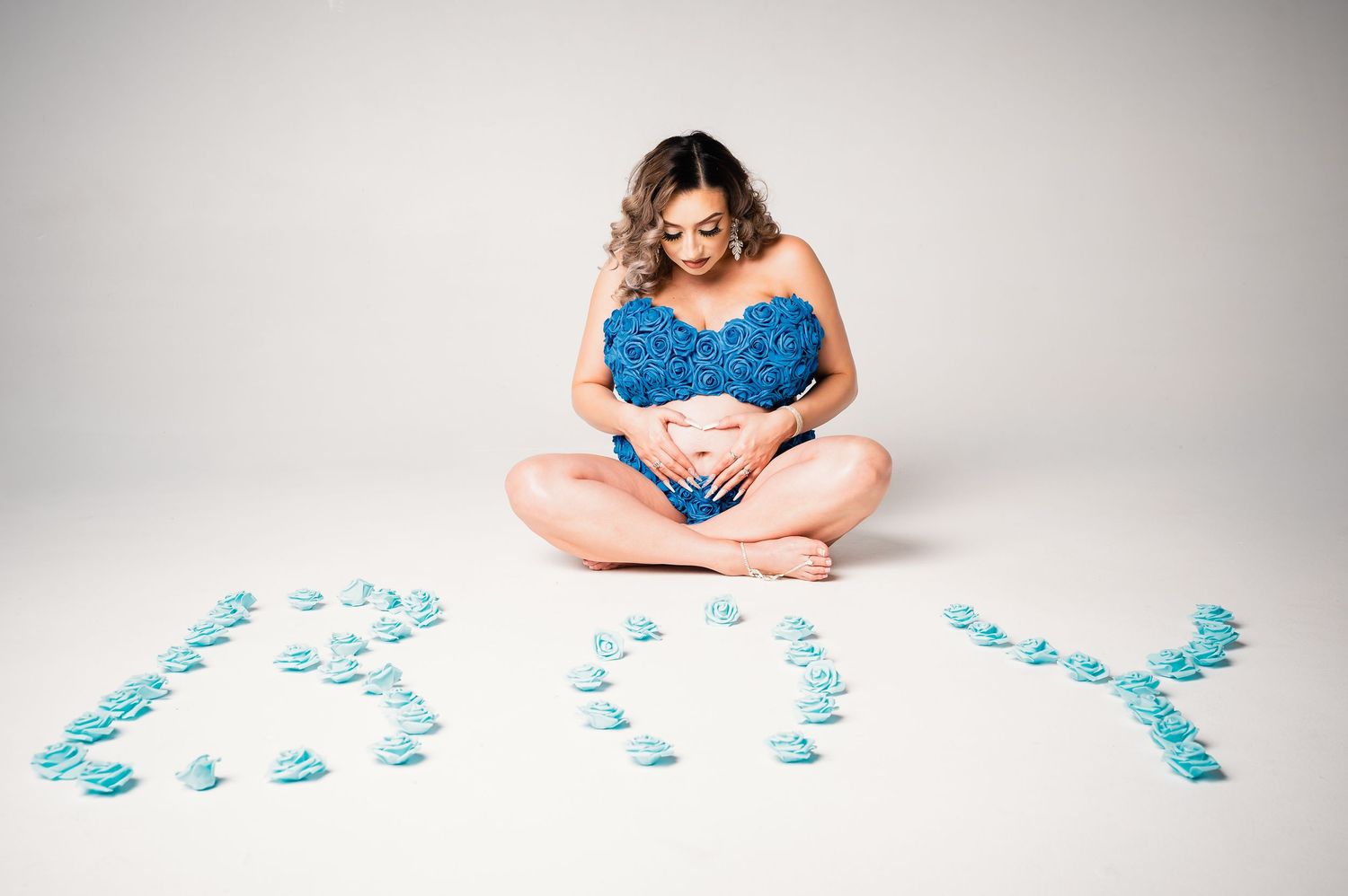 Pregnant woman in blue top sits cross-legged surrounded by blue roses spelling BOY on white background.