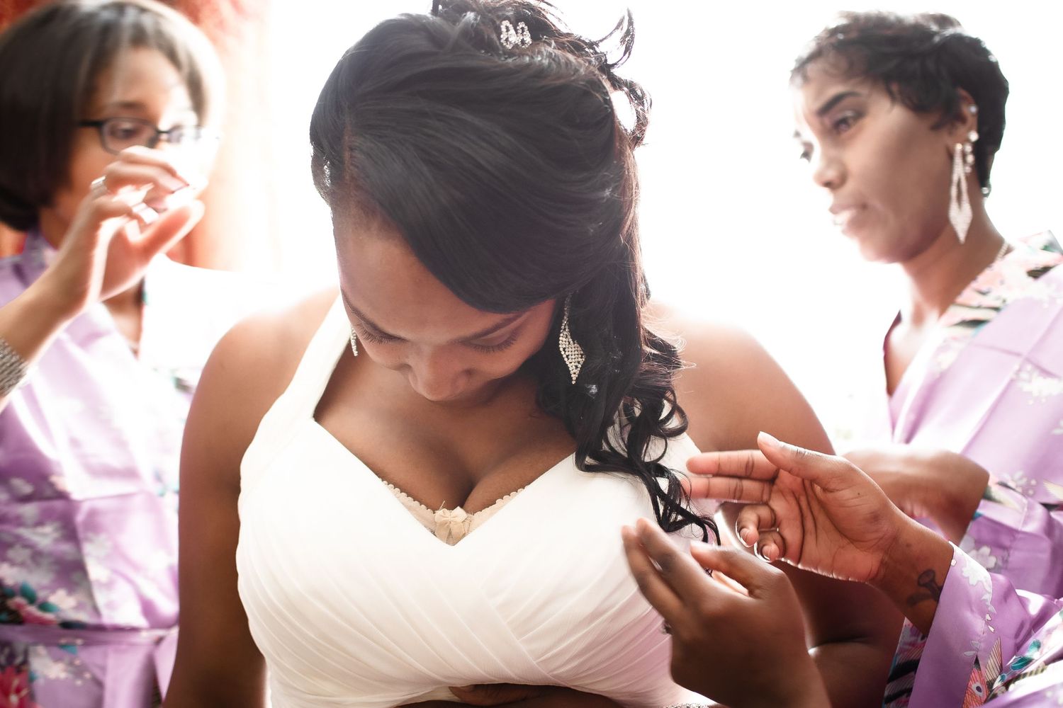 Bridesmaids in pink robes help bride with dress preparations before wedding ceremony.