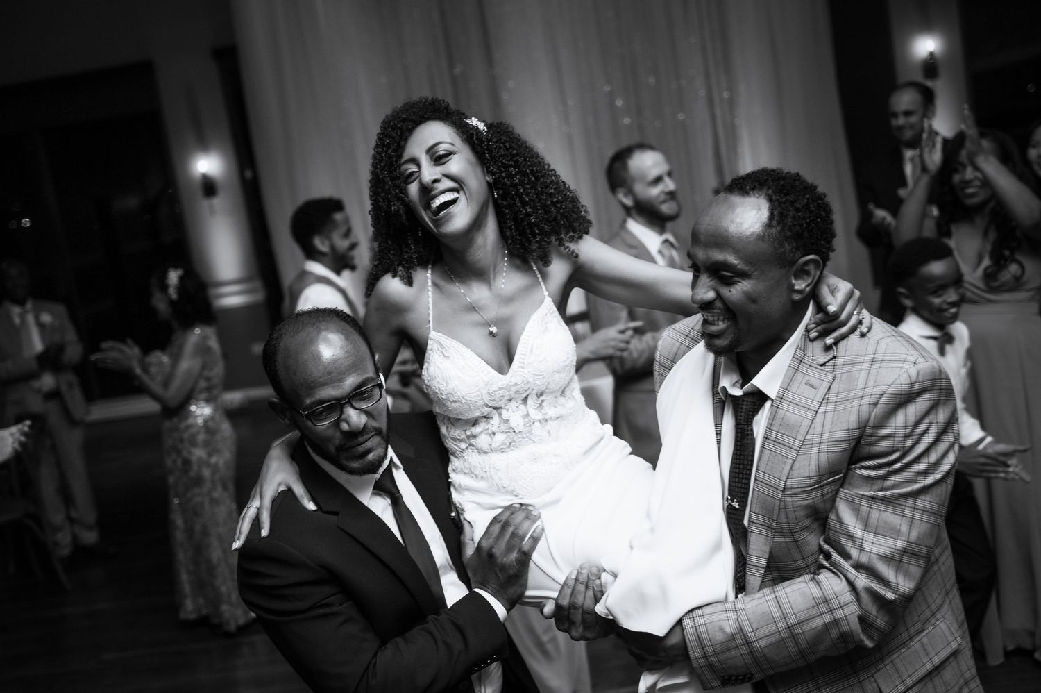 A joyful bride is lifted by two men during a lively black and white wedding reception dance.