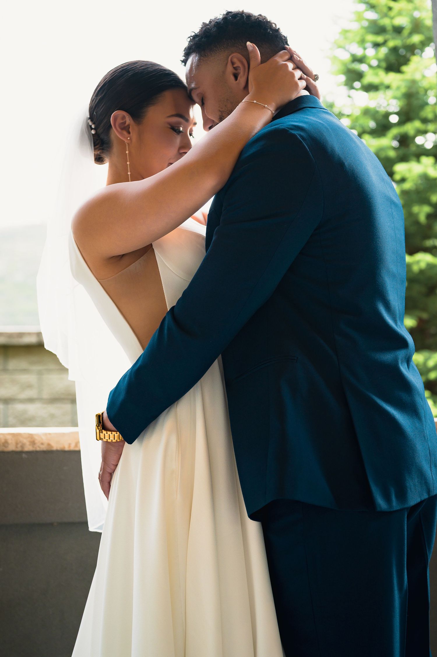 A bride and groom share an intimate embrace, she in a white gown with veil, he in a teal suit, outdoors.