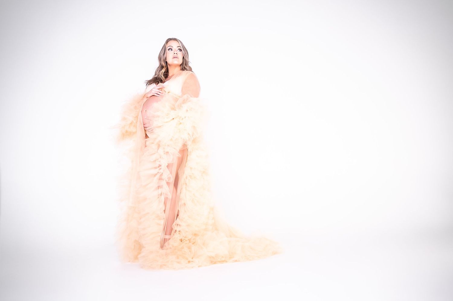 Pregnant woman posing in elegant peach feather maternity gown against white studio background.