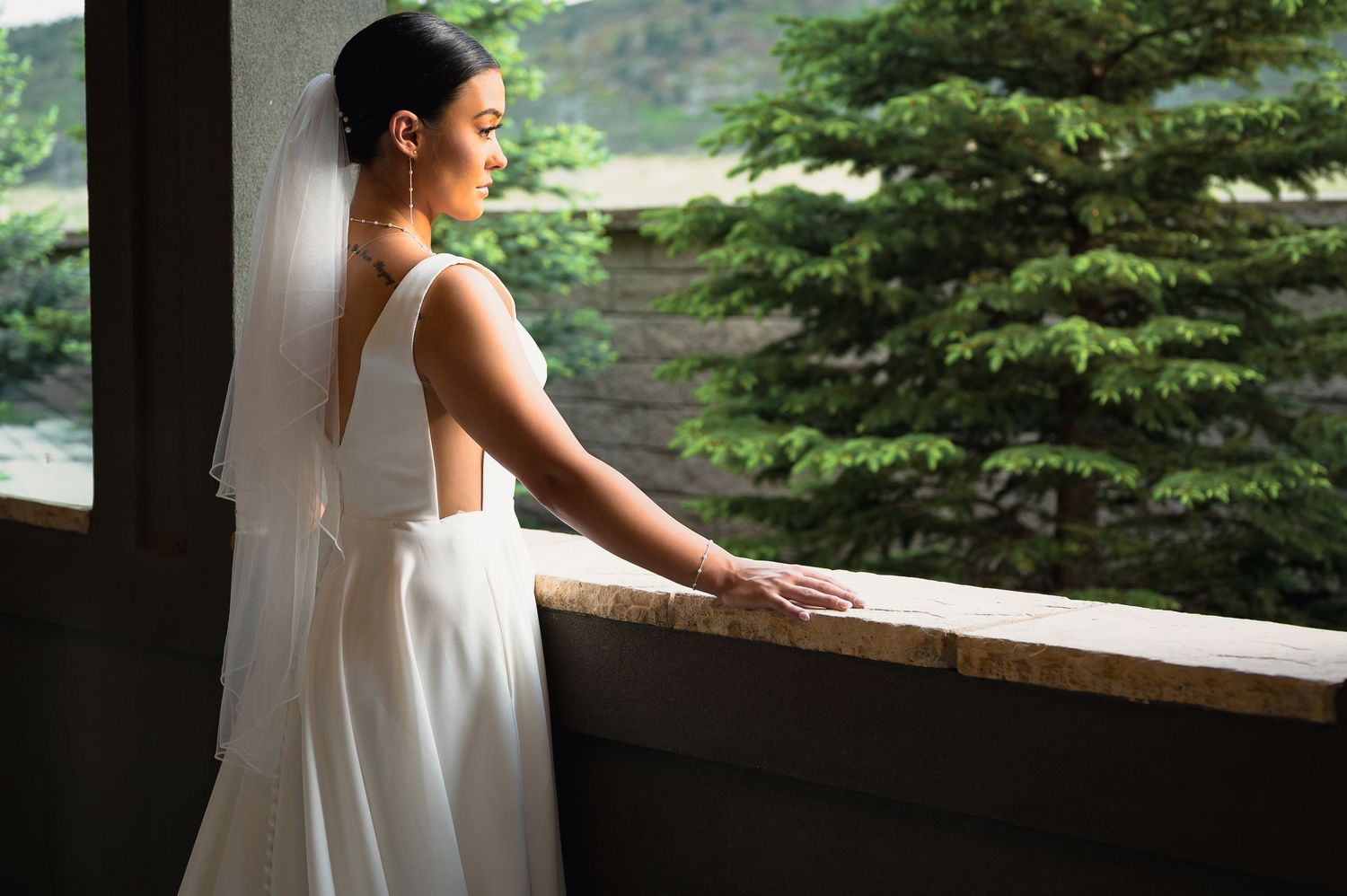 Elegant bride in a backless white gown and veil gazing out a window at lush green trees.
