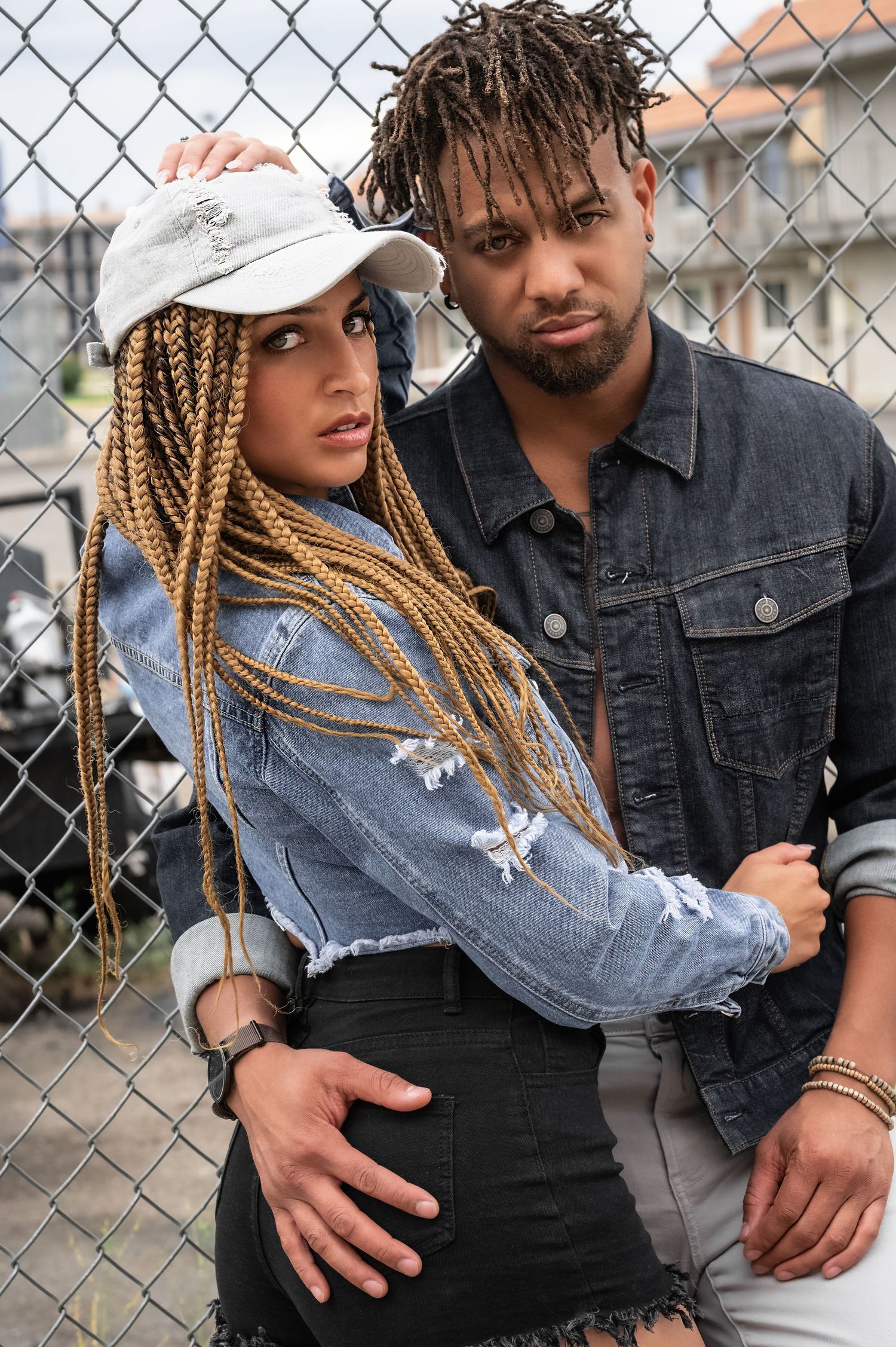 Stylish urban couple posing against a chain-link fence, woman with long blonde box braids.