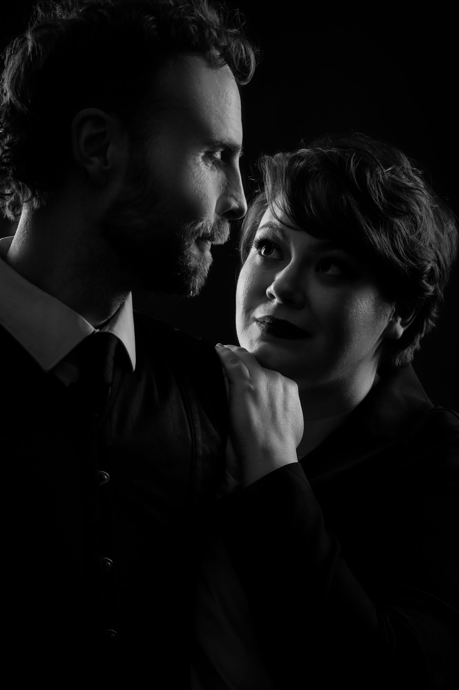 Dramatic black and white studio portrait of a bearded man and woman facing each other closely.