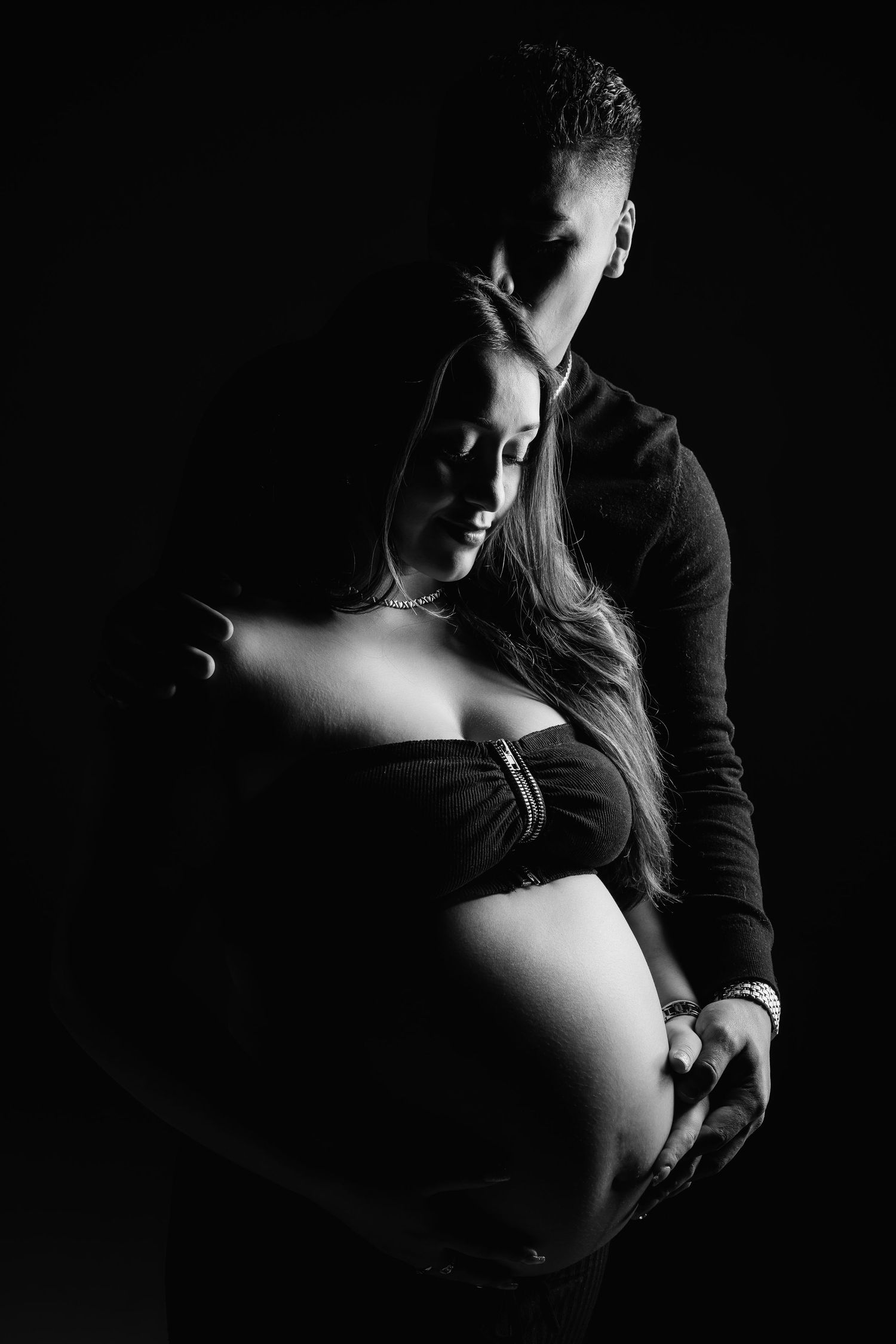 Black and white maternity photo of couple, man tenderly embracing pregnant woman from behind.
