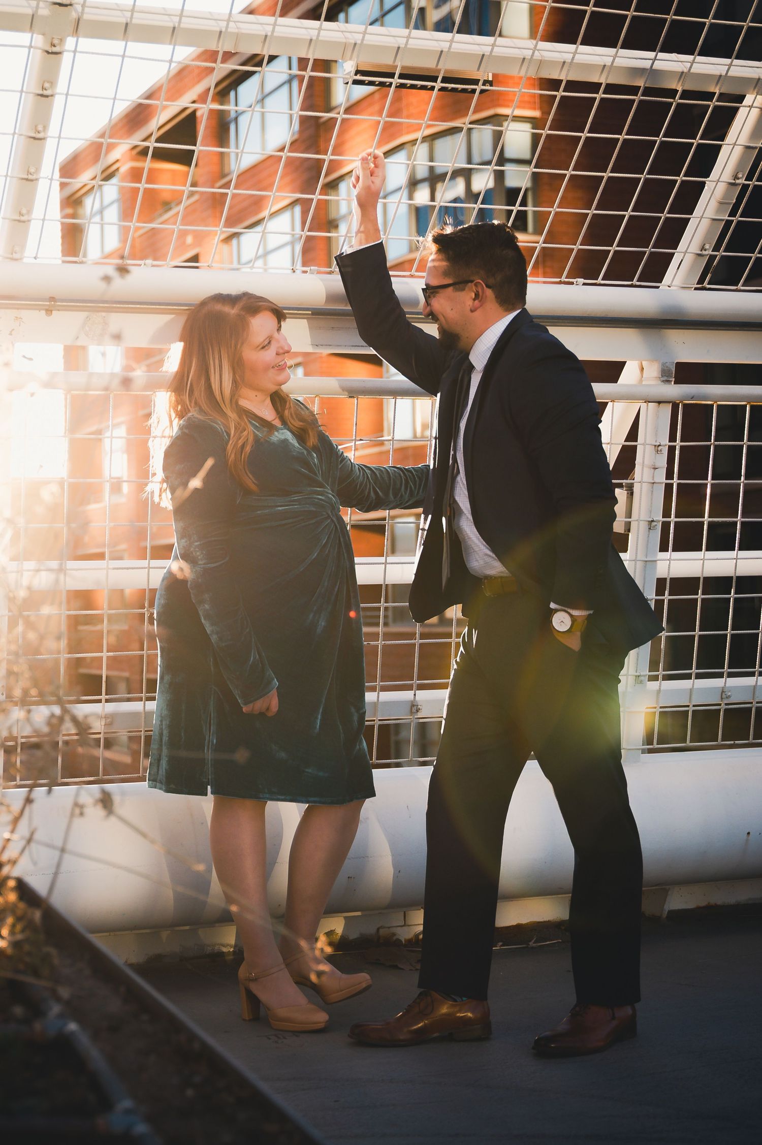 Pregnant couple dancing joyfully on a bridge bathed in warm golden sunset light.
