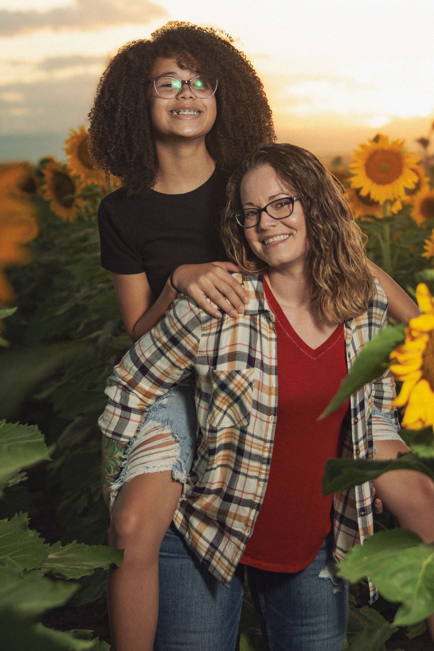 A woman giving a curly-haired girl a piggyback ride in a sunflower field at sunset.