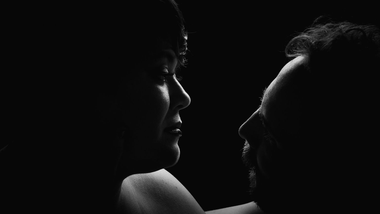 Black and white intimate portrait of couple facing each other in dramatic low-key lighting.