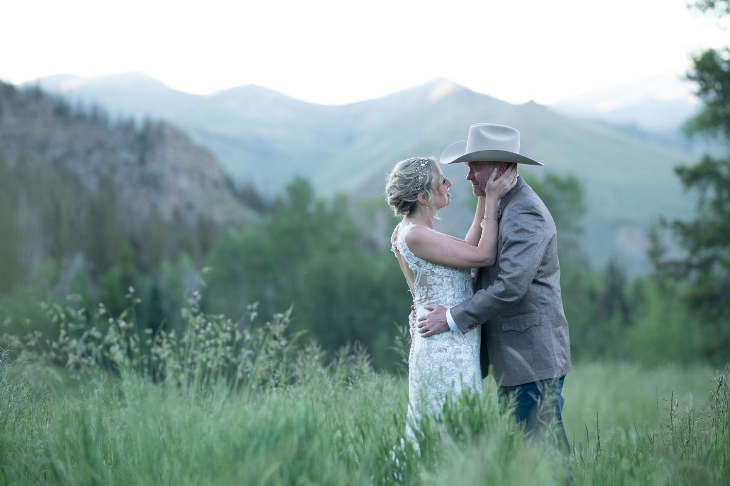 Bride and groom embrace romantically in a lush green mountain meadow at a rustic outdoor wedding.