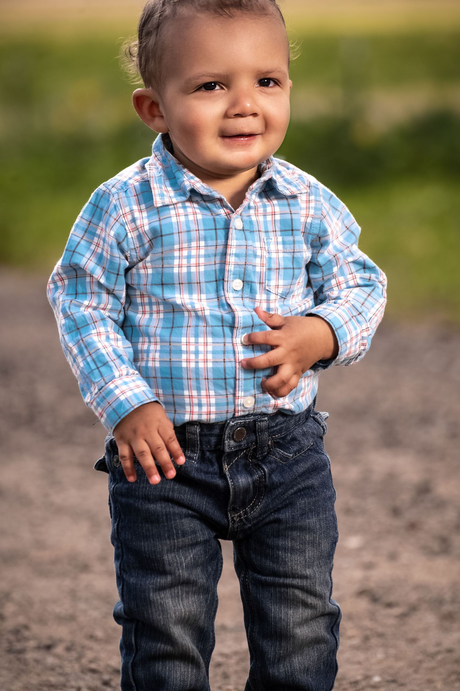 Smiling toddler boy in blue plaid shirt and dark jeans standing outdoors on a gravel path.