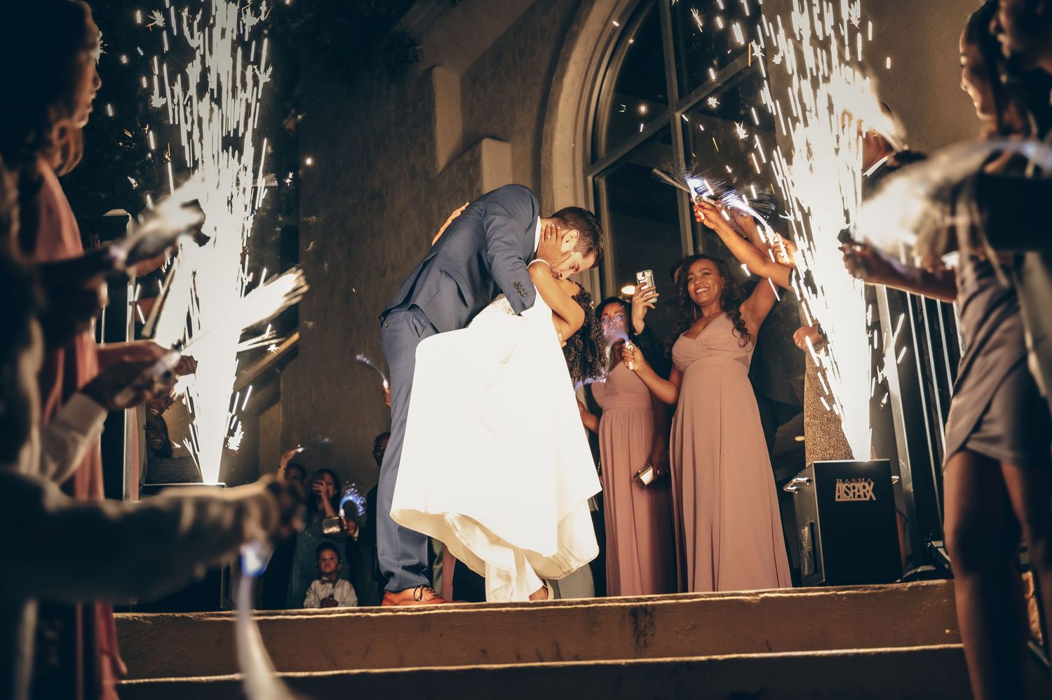 Bride and groom kiss on church steps surrounded by sparklers and bridesmaids in pink.