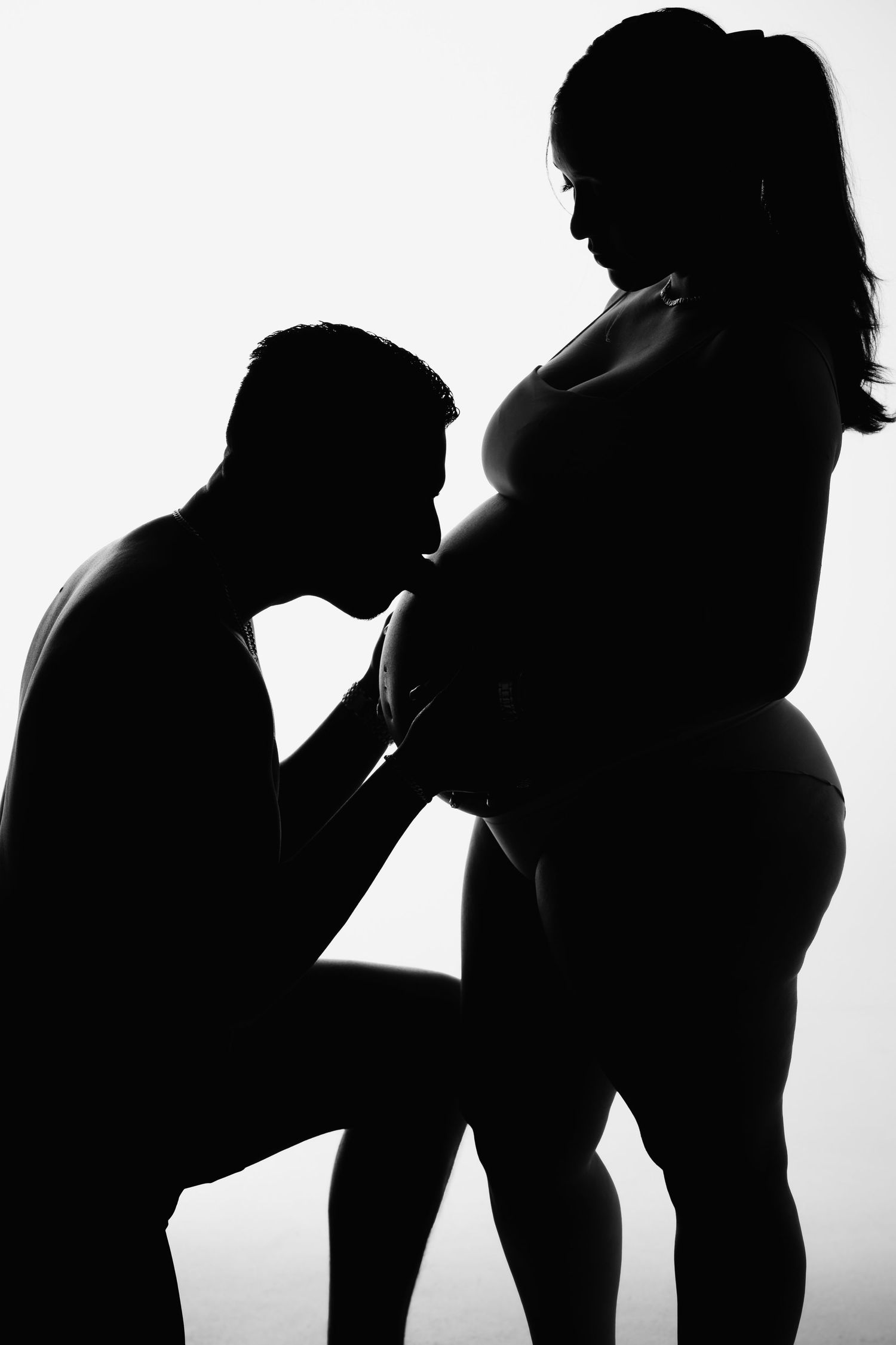 Silhouette of man kneeling to kiss pregnant woman's belly against bright white background.