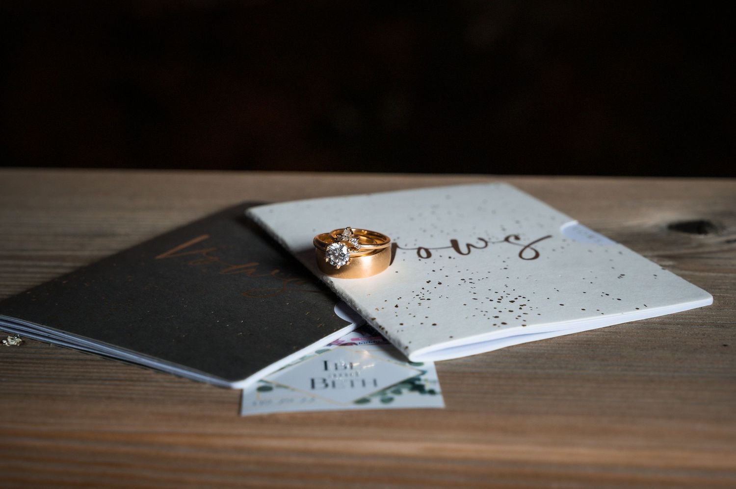 Gold diamond engagement ring resting on wedding vow books and cards on rustic wooden table.