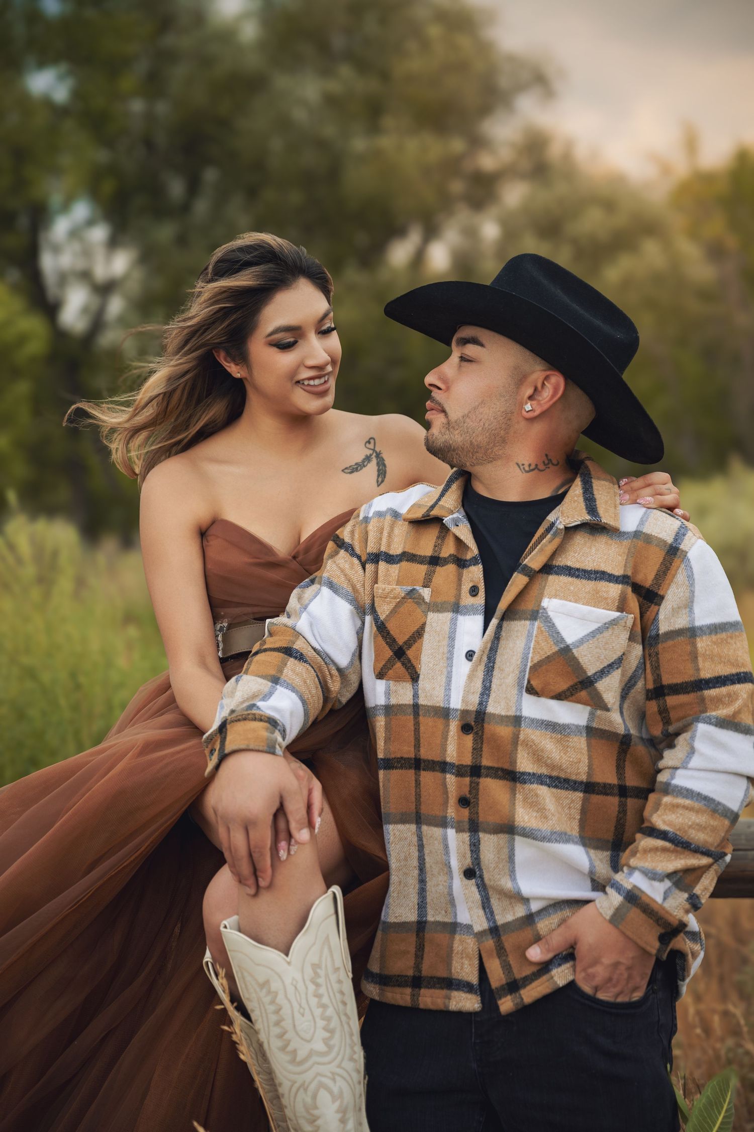 Couple posing outdoors in autumn field, woman in brown tulle dress with cowboy boots, man in plaid shirt and black hat.