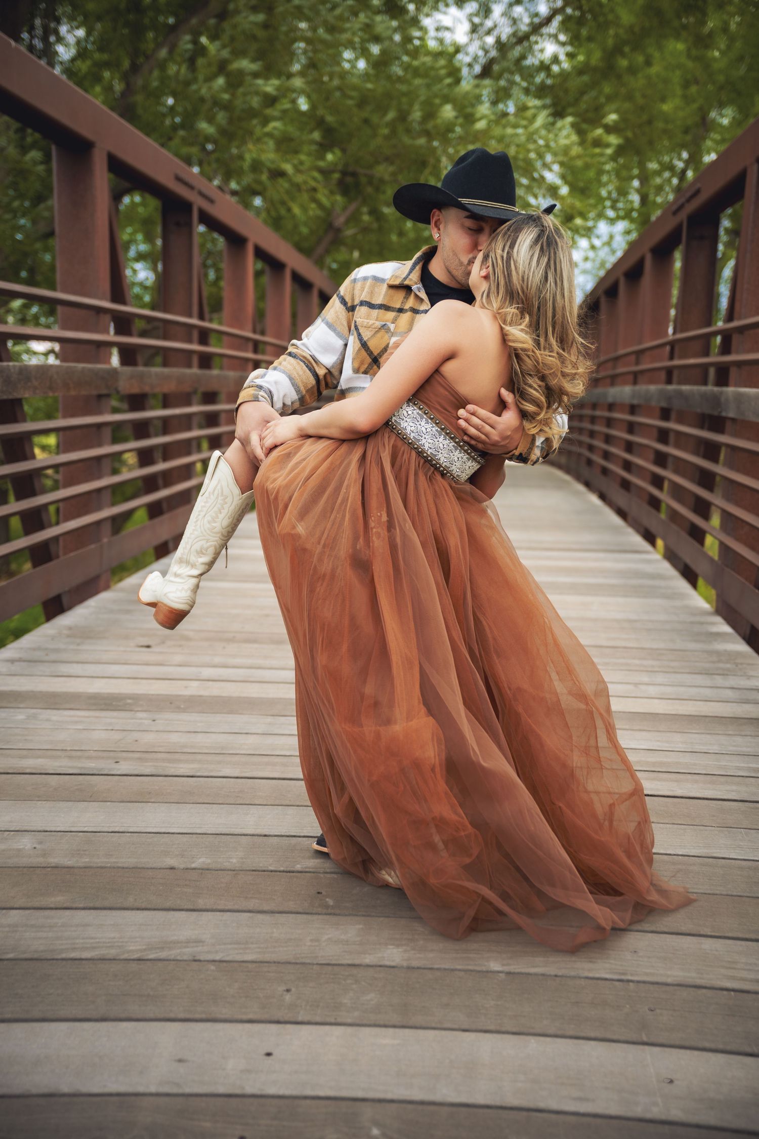 Romantic couple kissing on a wooden bridge, woman in flowing brown gown and white boots, man in cowboy hat lifting her.