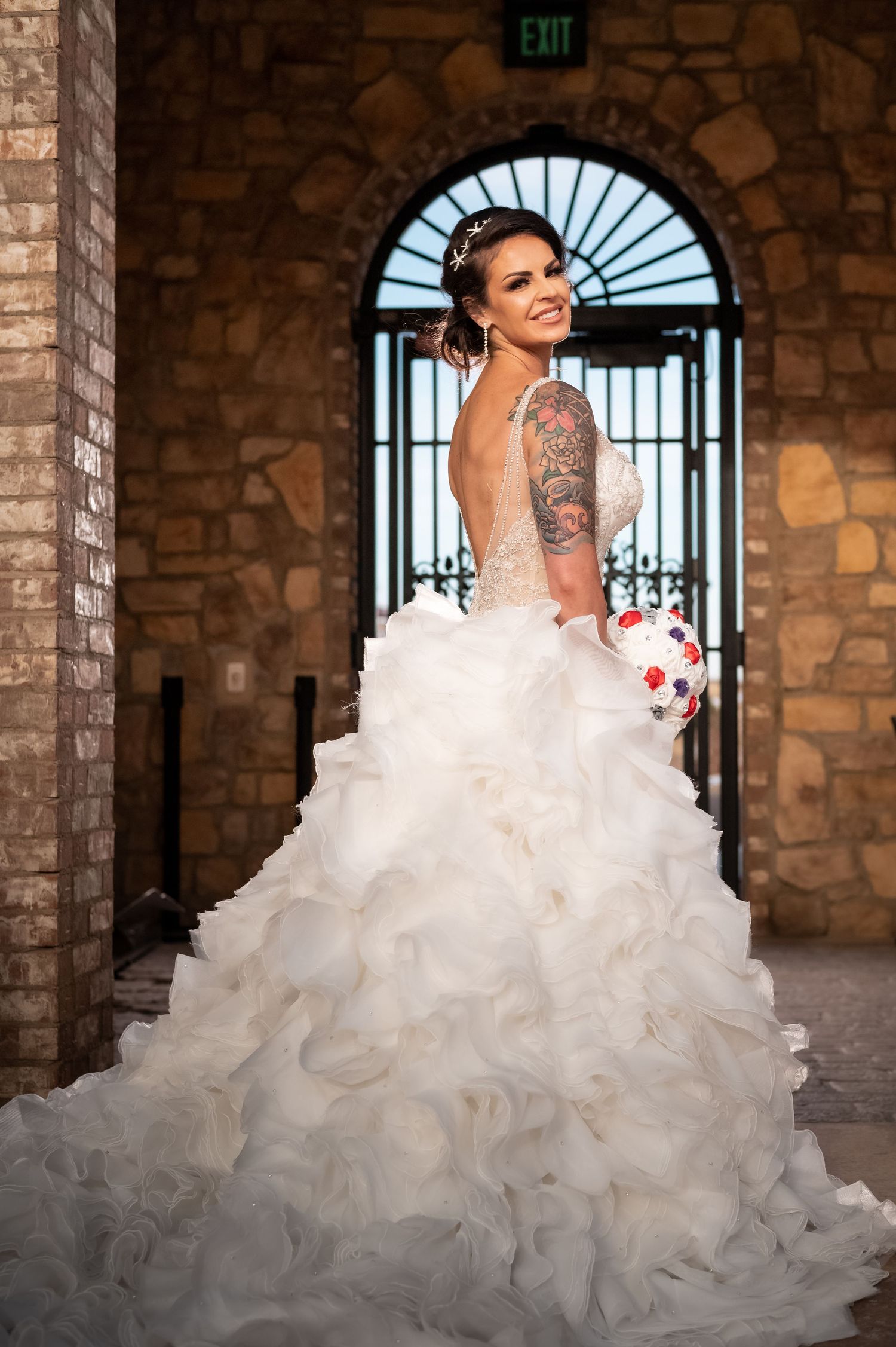 Smiling bride in backless ruffled ball gown with tattoo sleeve holding bouquet in stone venue.