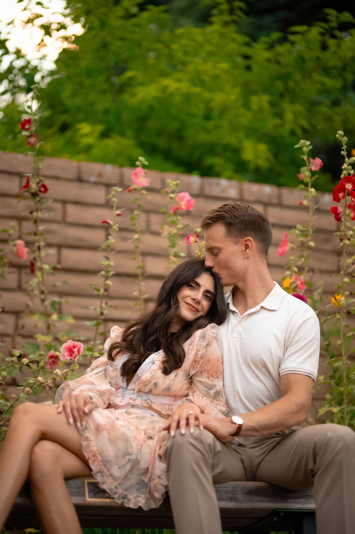 Couple sitting on a bench surrounded by pink hollyhock flowers, man kissing woman's head in a lush garden.