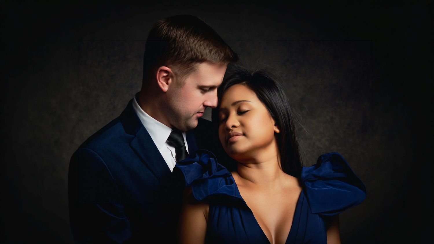 Elegant couple in blue formal wear sharing an intimate moment against a dark backdrop.