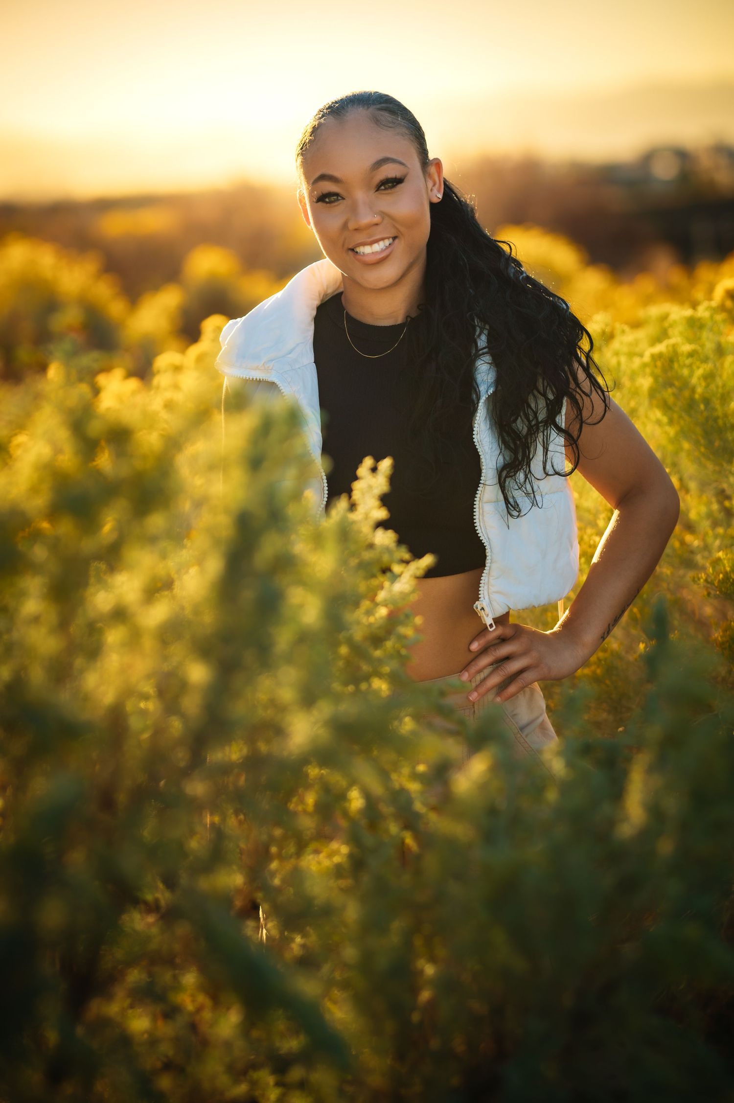 Smiling young Black woman in black crop top standing in golden field at sunset.