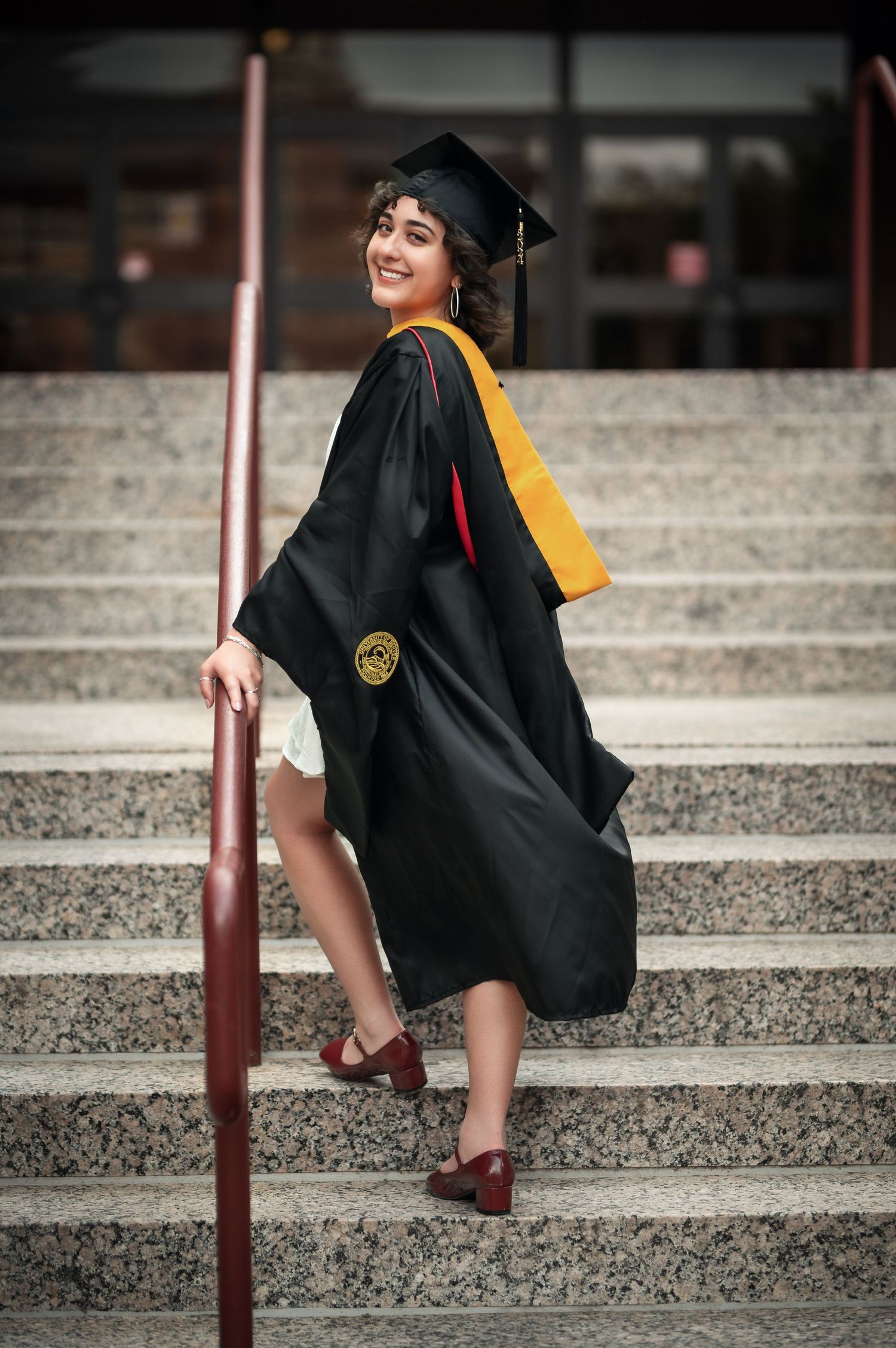 A smiling graduate in black and gold cap and gown poses on outdoor university steps.