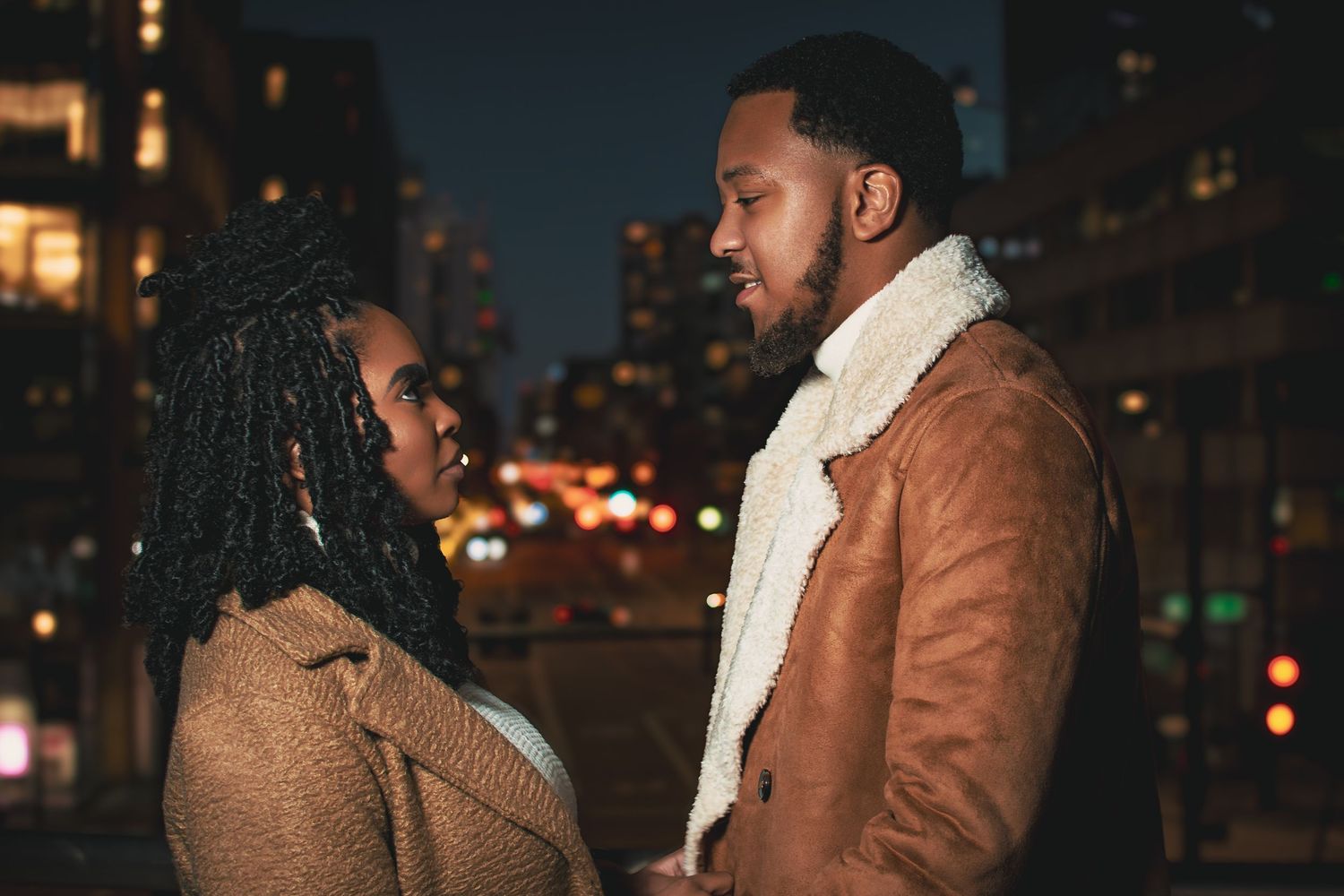 Romantic couple in winter coats facing each other against a blurred city lights backdrop at night.
