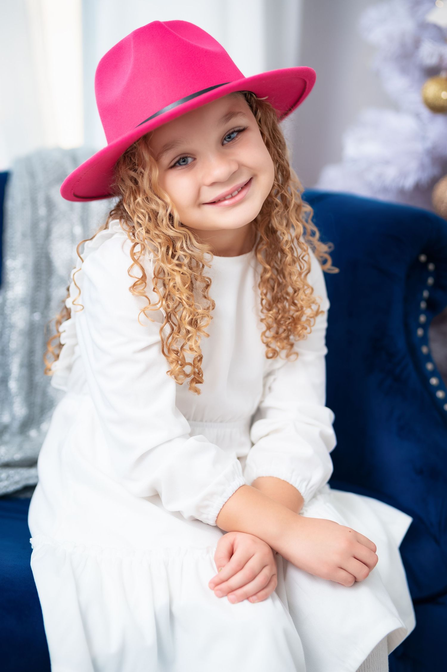 Smiling girl with curly blonde hair wearing pink hat and white dress sitting on blue chair.
