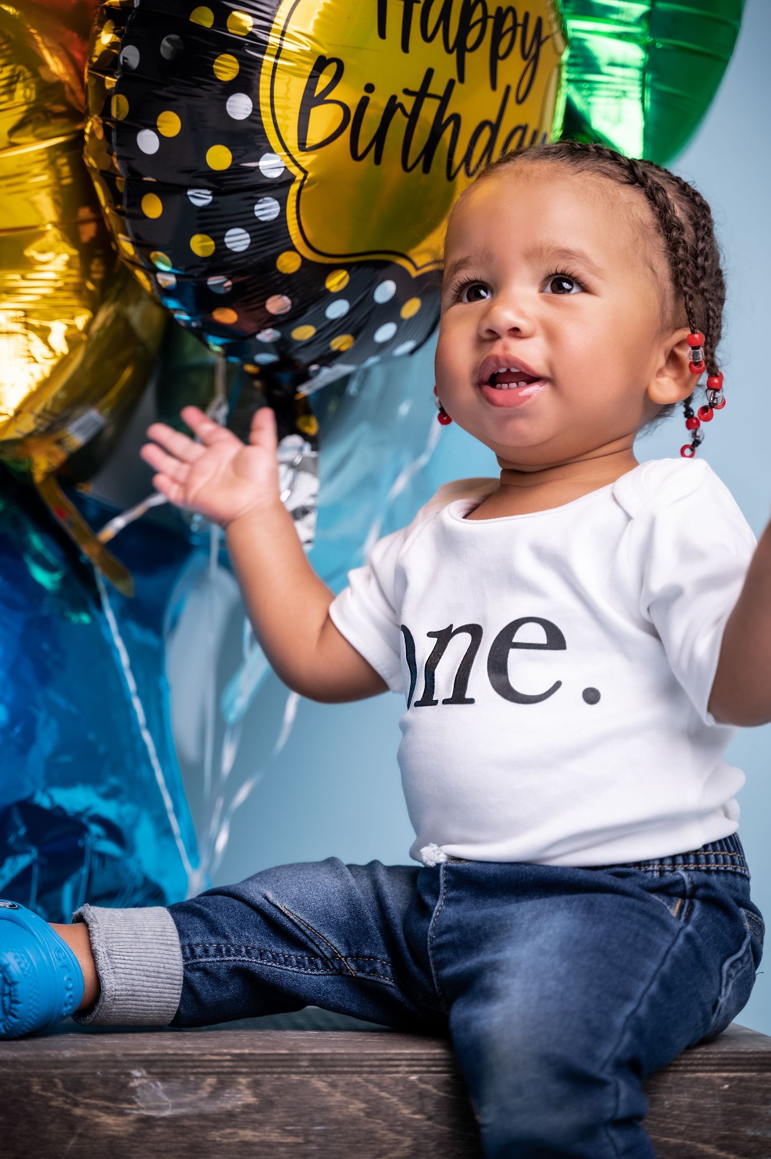 Baby celebrating first birthday wearing 'one' onesie surrounded by colorful balloons.