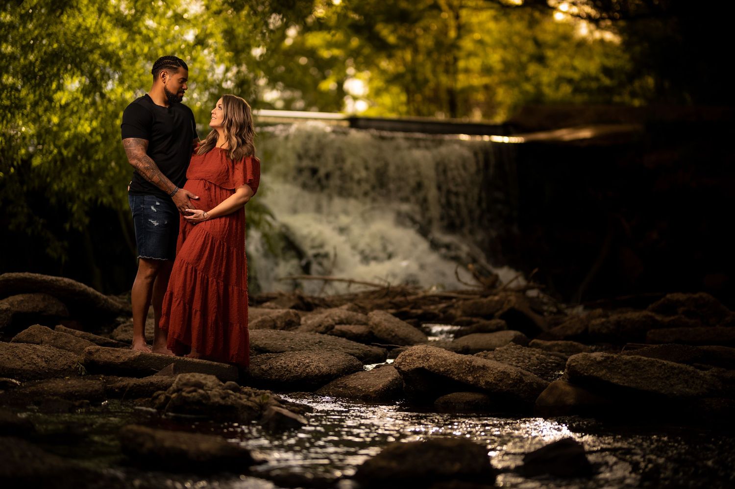 Couple expecting a baby standing on rocks before a waterfall in a lush forest setting.