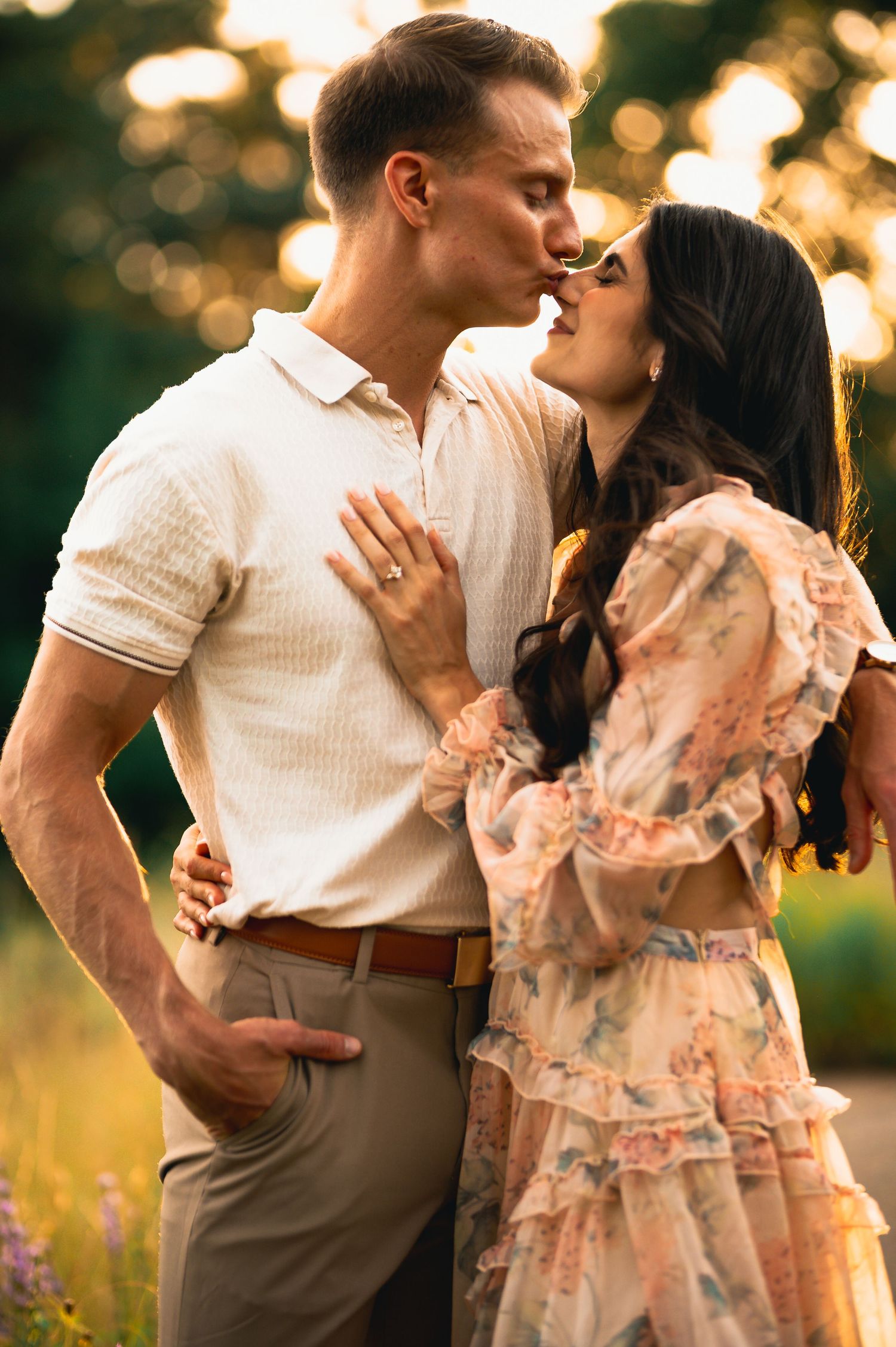 Couple sharing a romantic kiss outdoors at golden hour, woman in floral dress touching man's chest.
