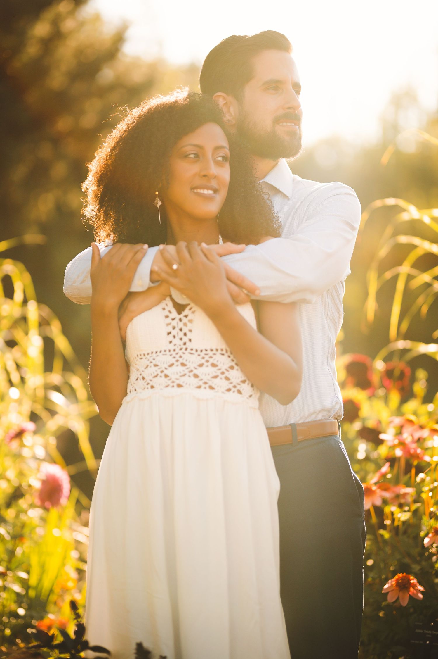 Interracial couple embracing in sunlit garden surrounded by colorful blooming flowers.
