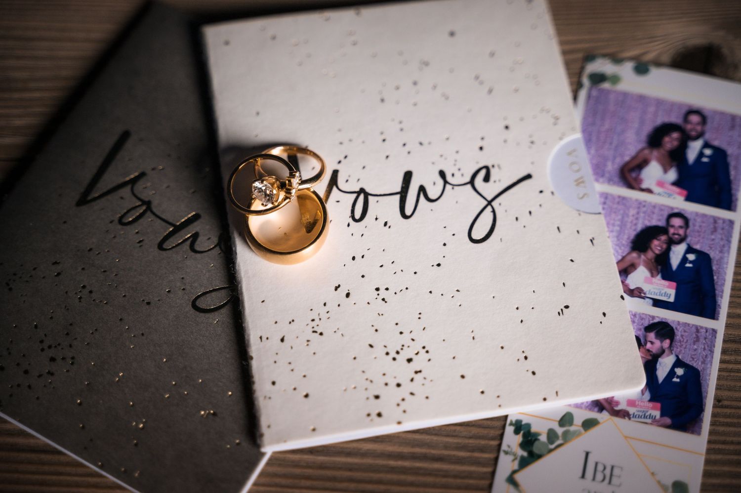 Gold wedding rings resting on white vow book with photo booth pictures on wooden table.