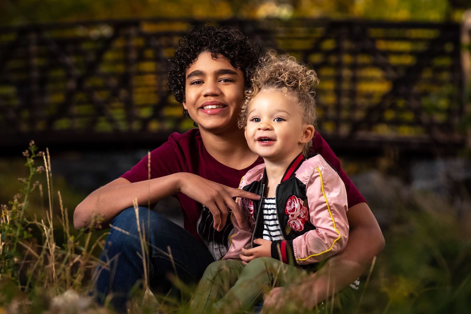 Two siblings smiling outdoors in a lush green field near a wooden bridge.