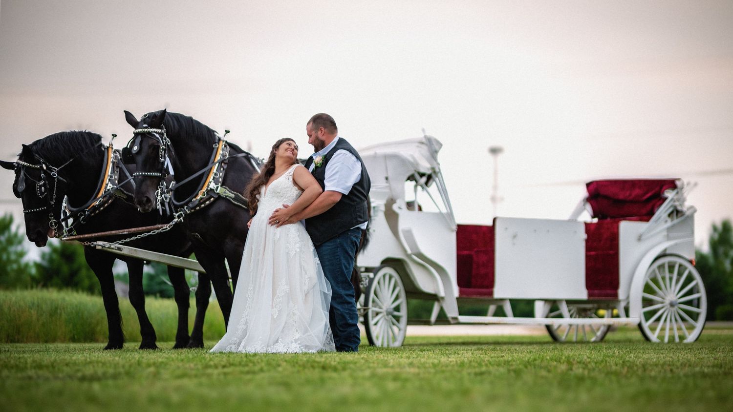 Bride and groom share a romantic kiss beside black horses and a white carriage at their wedding.