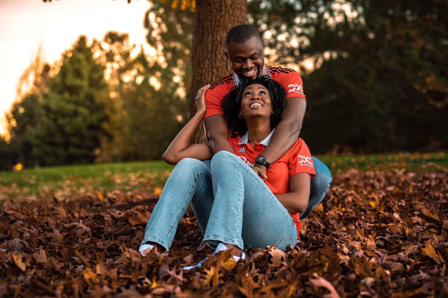 Happy couple sitting in autumn leaves against tree trunk in golden sunset light.