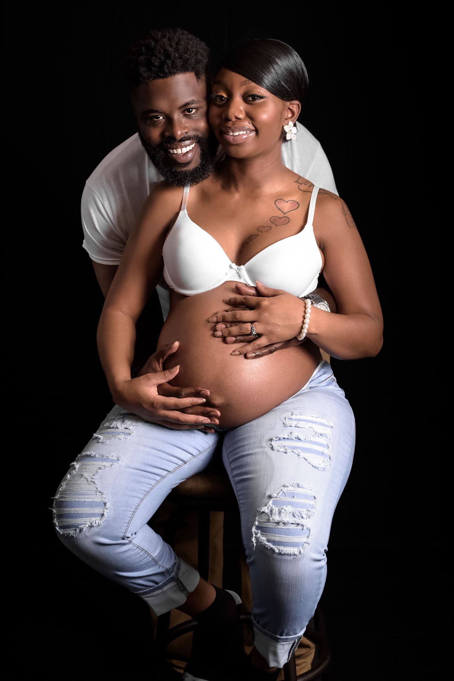 Smiling African American couple posing together, pregnant woman in white bra and jeans on black background.