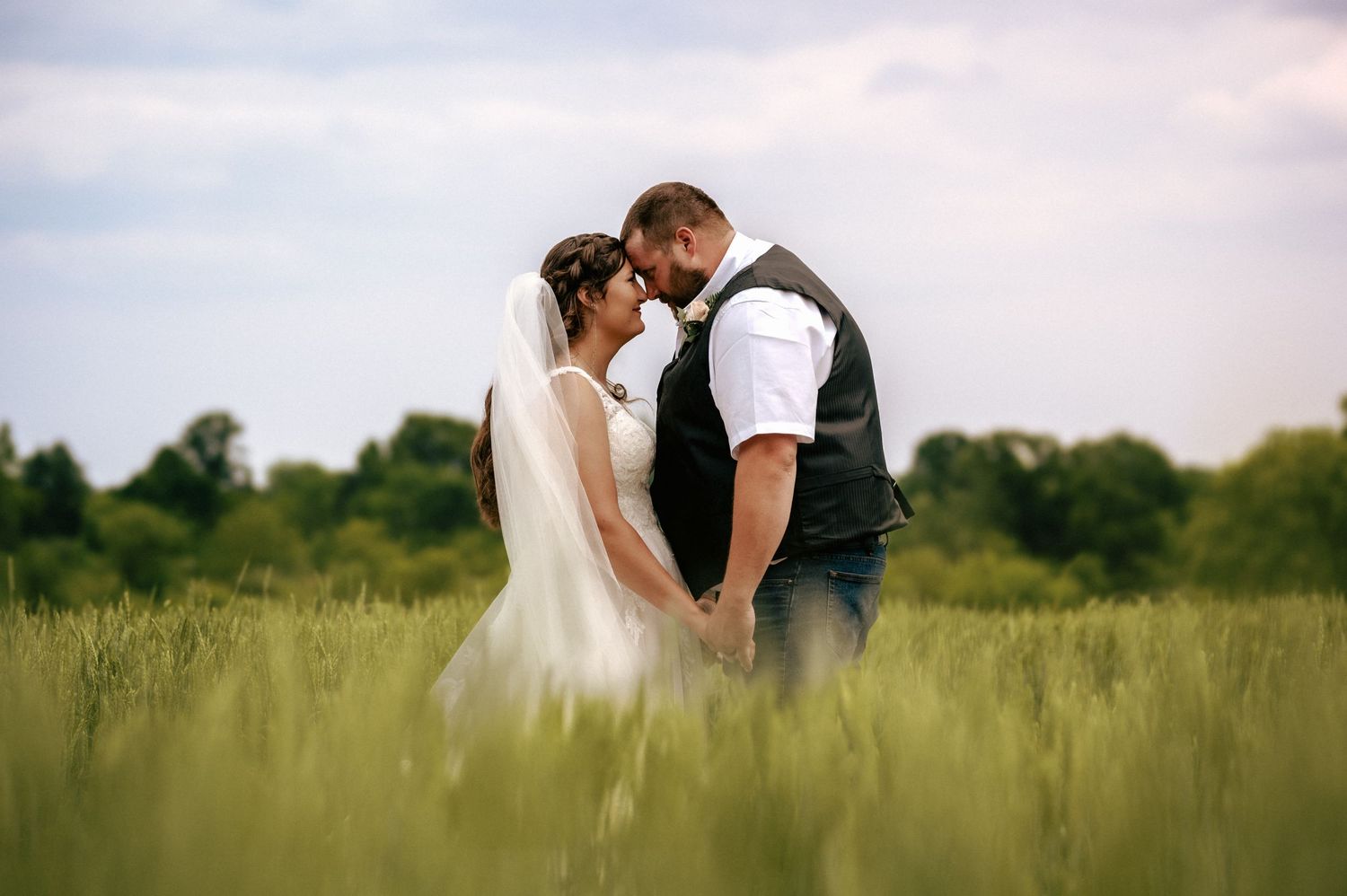 Bride and groom holding hands in lush green field under cloudy sky on wedding day.