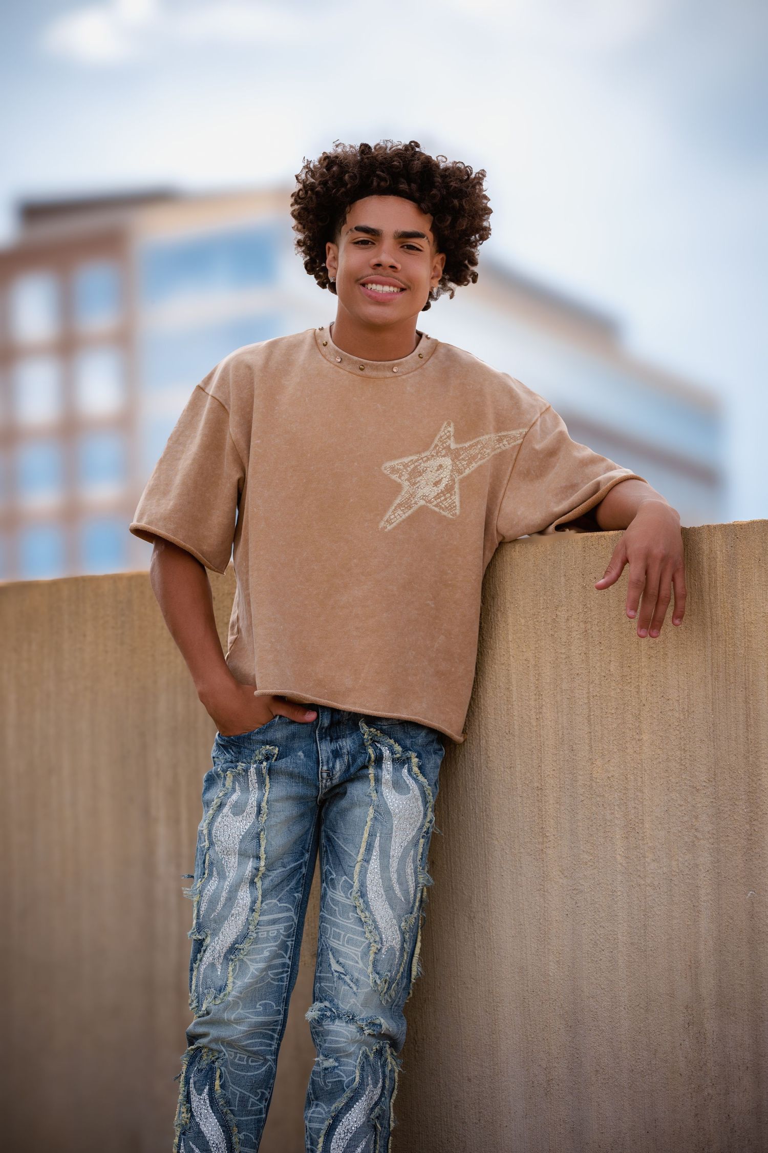 A smiling teen with curly hair leans against a wall in a tan graphic tee and jeans.