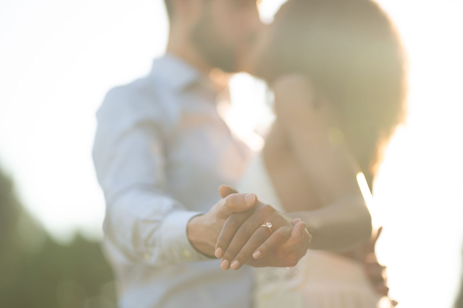 Romantic backlit photo of couple holding hands and kissing at sunset, engagement ring visible on woman's hand.
